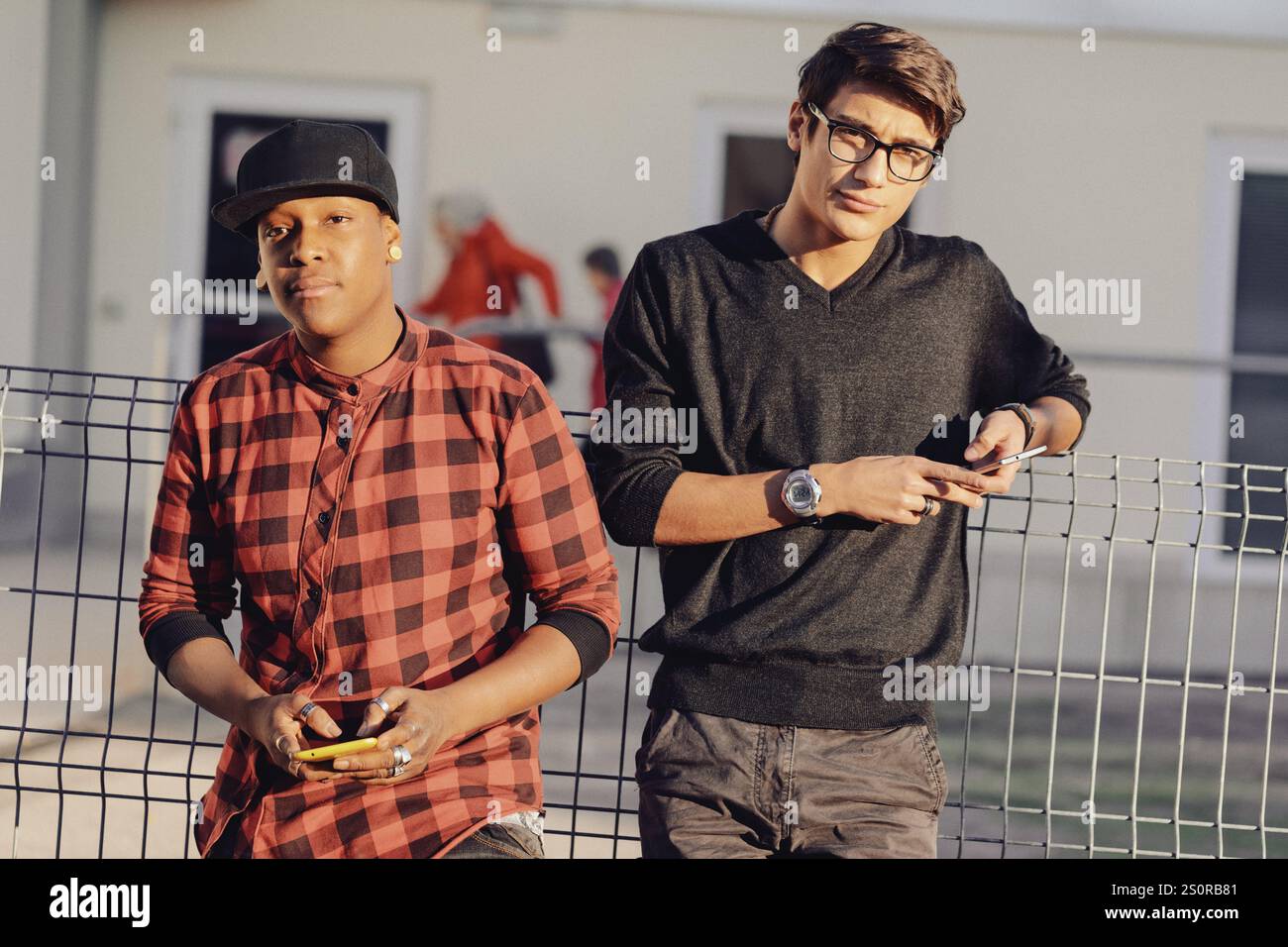 Deux jeunes hommes adultes penchés décontractés contre une clôture métallique, pris dans leurs smartphones tout en profitant d'un moment de détente à l'extérieur dans un décor urbain Banque D'Images