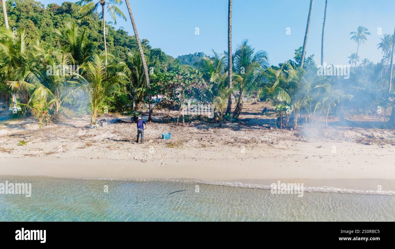 Koh Kood Thaïlande 20 décembre 2024, les gens brûlent des déchets sur la plage, la lumière du soleil filtre à travers de grands palmiers sur Koh Kood, comme un personnage se tient près du bord de l'eau tandis que la fumée dérive d'un endroit voisin Banque D'Images