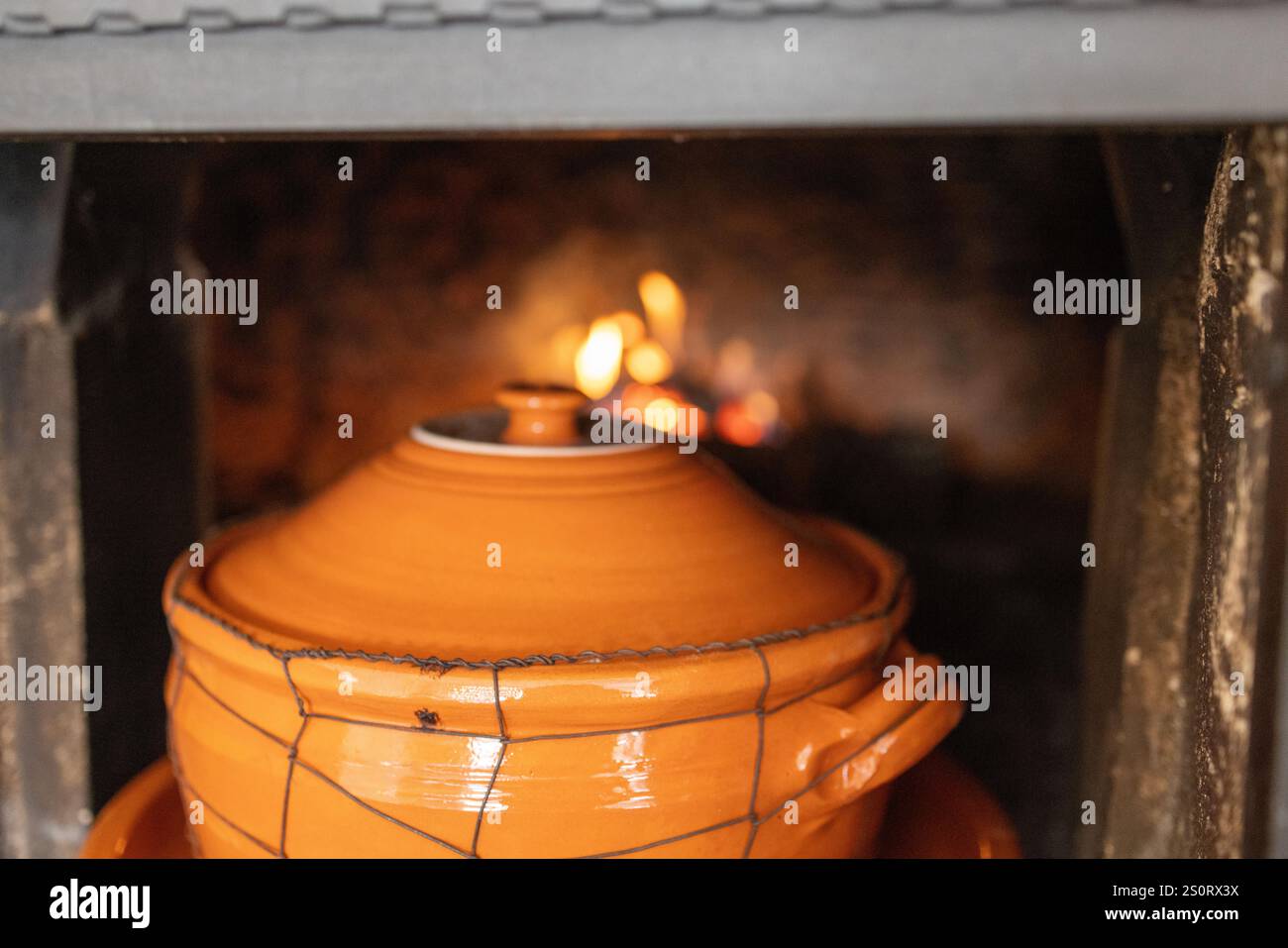 Gros plan d'un pot en argile placé dans un four à bois, avec des flammes incandescentes en arrière-plan. La scène capture le charme rustique de la cuisine traditionnelle Banque D'Images