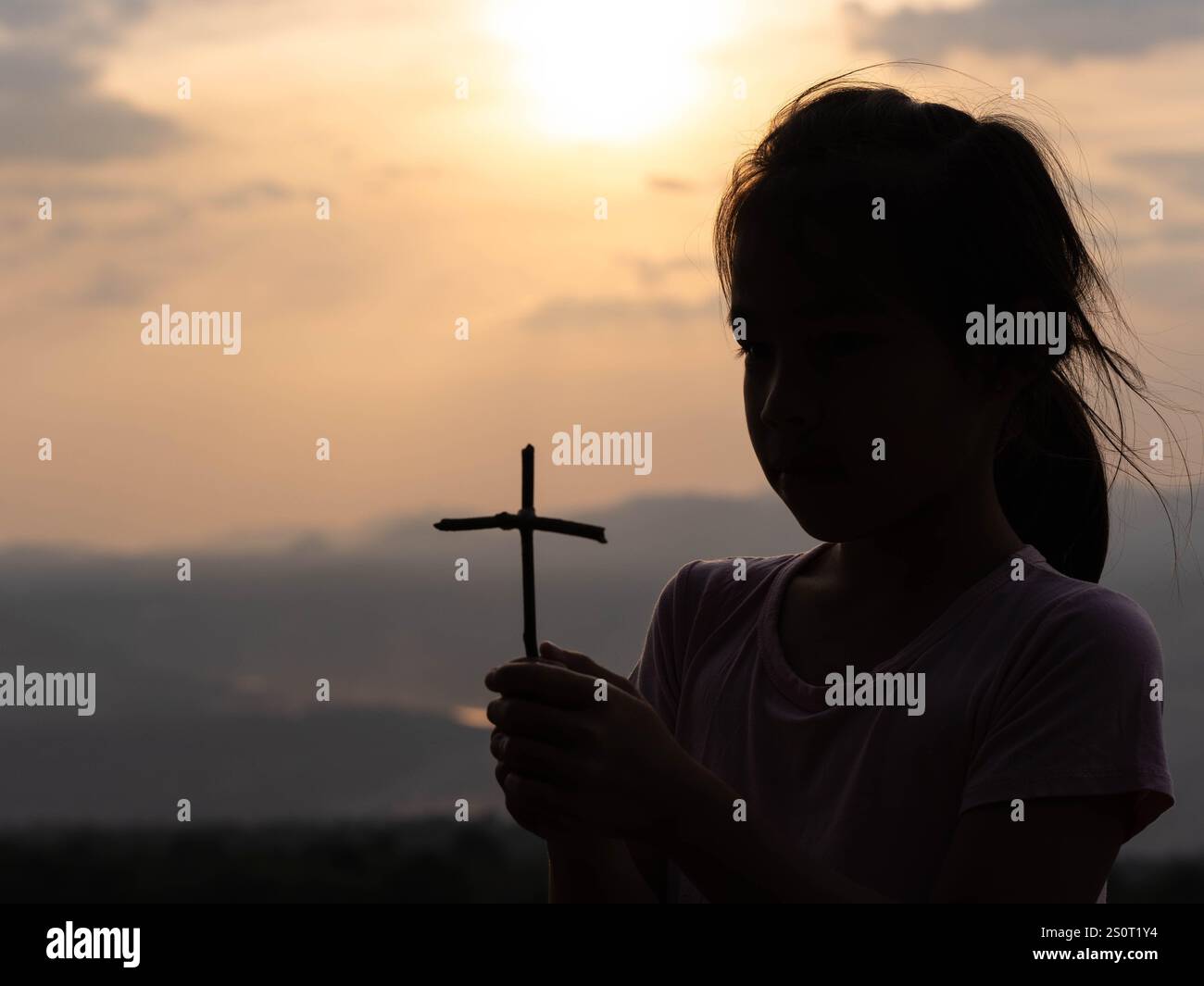 Silhouette d'une petite fille mignonne tenant une croix priant seule dans un parc contre un coucher de soleil chaud. Banque D'Images