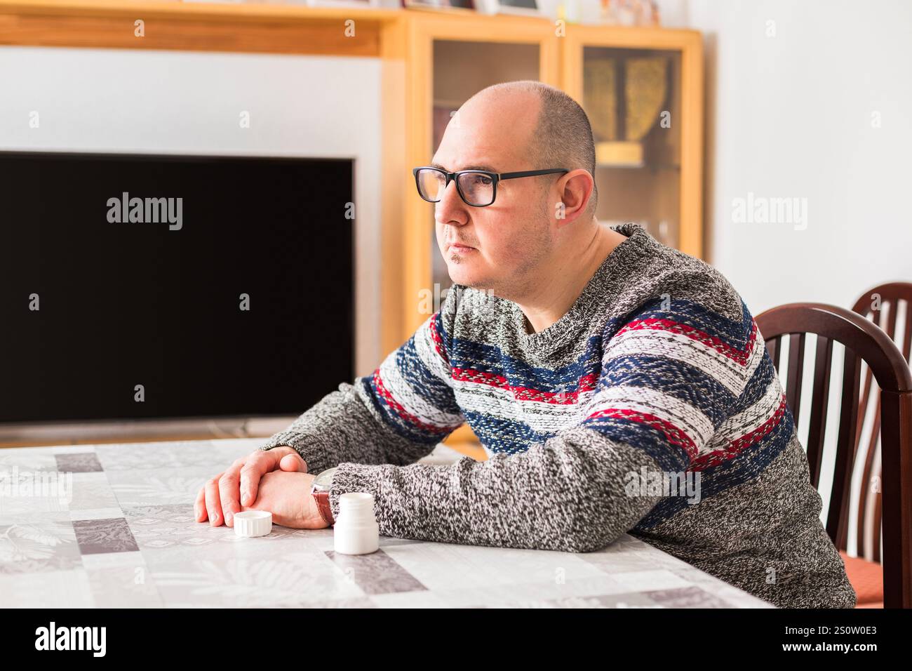 Un homme adulte chauve assis à une table à côté d'une bouteille de médicament avec un regard triste et inquiet vers l'horizon. Banque D'Images