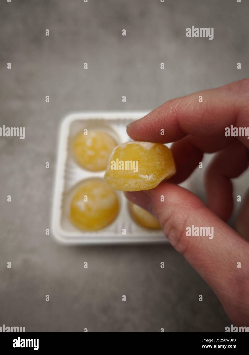 Ramasser à la main un seul mochi à la mangue jaune dans un plateau en plastique, prêt à profiter d'une friandise sucrée Banque D'Images