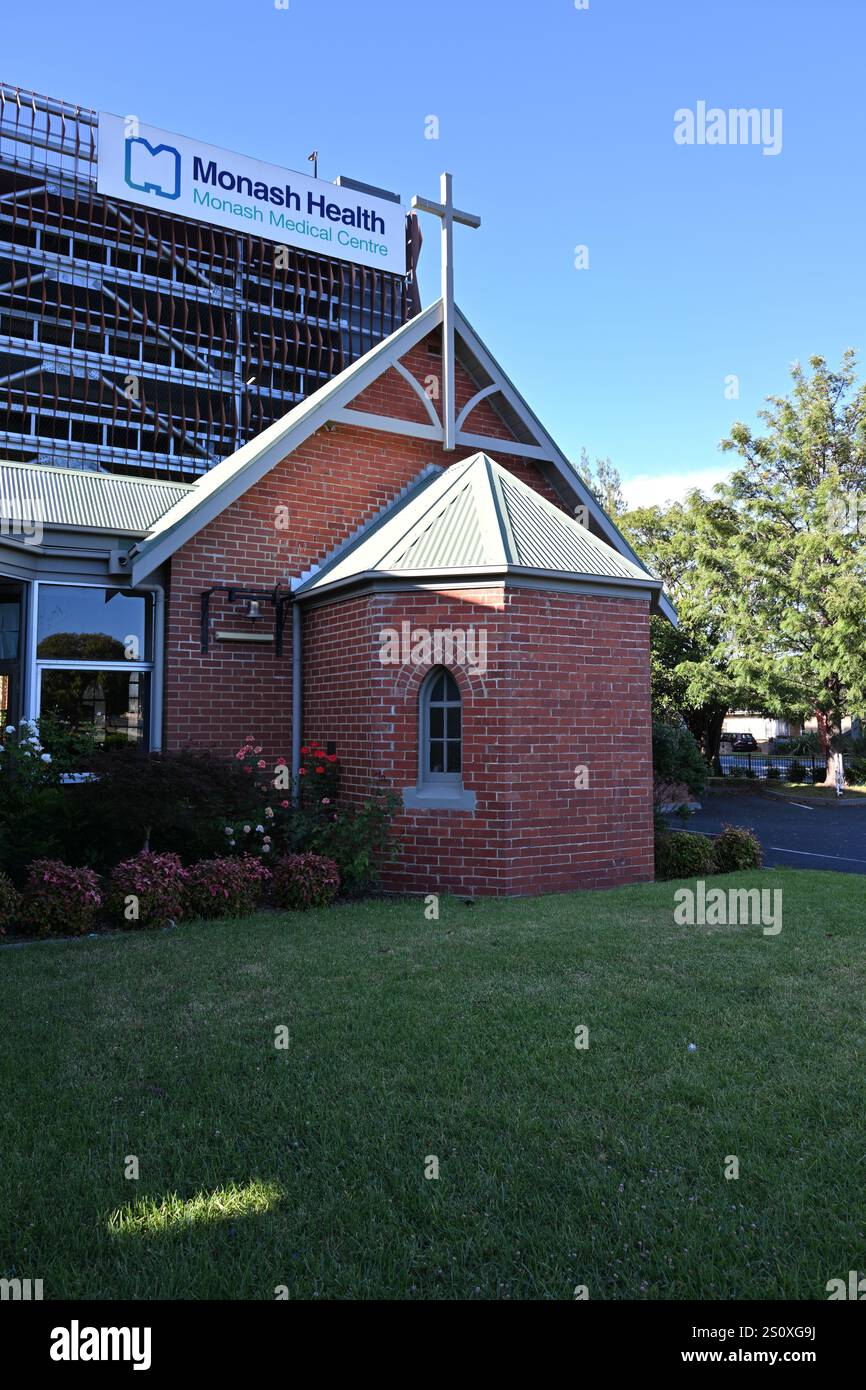 Modeste église anglicane de brique rouge All Saints avec le grand parking du Monash Medical Centre qui se profile à l'arrière-plan Banque D'Images