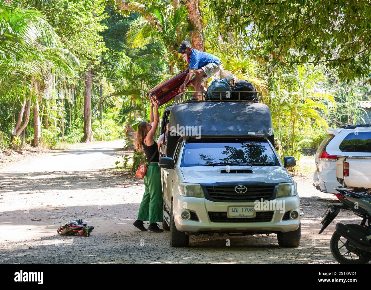 Un chauffeur de taxi local qui se tient sur le toit de son taxi avec les bagages d'autres passagers est vu prendre les bagages d'une voyageuse occidentale, dans un parking d'une station balnéaire de jardin, à Khlong Prao Beach, sur l'île de Koh Chang. Koh Chang, situé dans la province thaïlandaise de Trat, est une destination idéale pour les occidentaux qui cherchent à échapper à l'hiver. Décembre est l'un des meilleurs moments pour visiter, car le temps est sec et ensoleillé, ce qui le rend parfait pour se détendre sur la plage et explorer la beauté naturelle de l'île. Koh Chang a conservé une grande partie de son charme local. Alors que des équipements modernes sont disponibles, l'île a éviter Banque D'Images