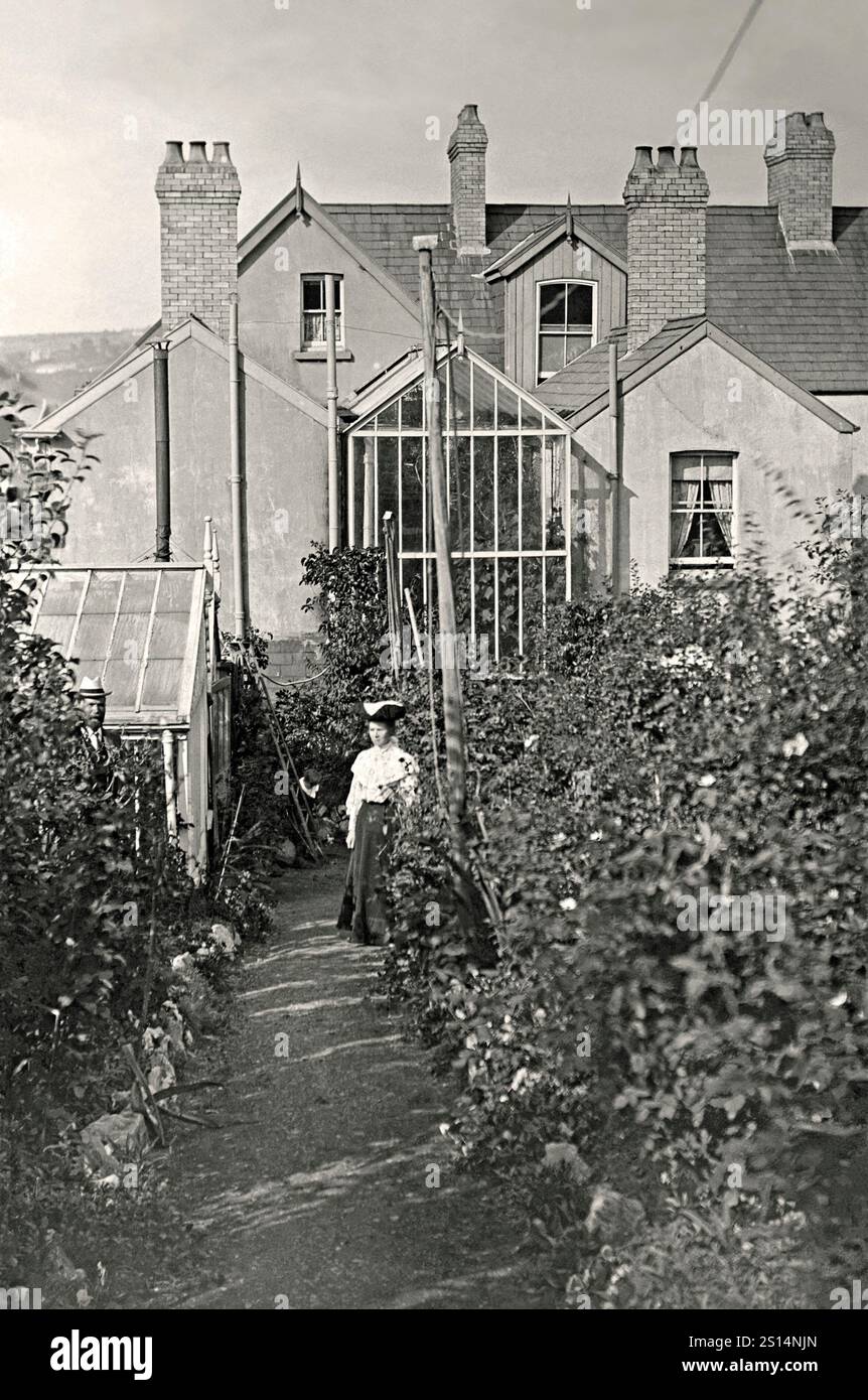 Une très grande serre construite dans le retour latéral d'une modeste maison mitoyenne à Porthcawl, Bridgend, pays de Galles vers 1900. Cette impressionnante structure vitrée remplit tout l’espace dans le retour latéral entre la maison et son voisin. Les propriétaires posent sur le chemin du jardin arrière. La maison de verre aurait permis de cultiver des plantes tendres qui pourraient mûrir à une assez grande hauteur - mais nettoyer le verre n'aurait pas été facile. Ceci est tiré d'une petite photographie victorienne. Il aura l'air doux si utilisé à une taille trop grande - une photographie vintage des années 1800/1900. Banque D'Images