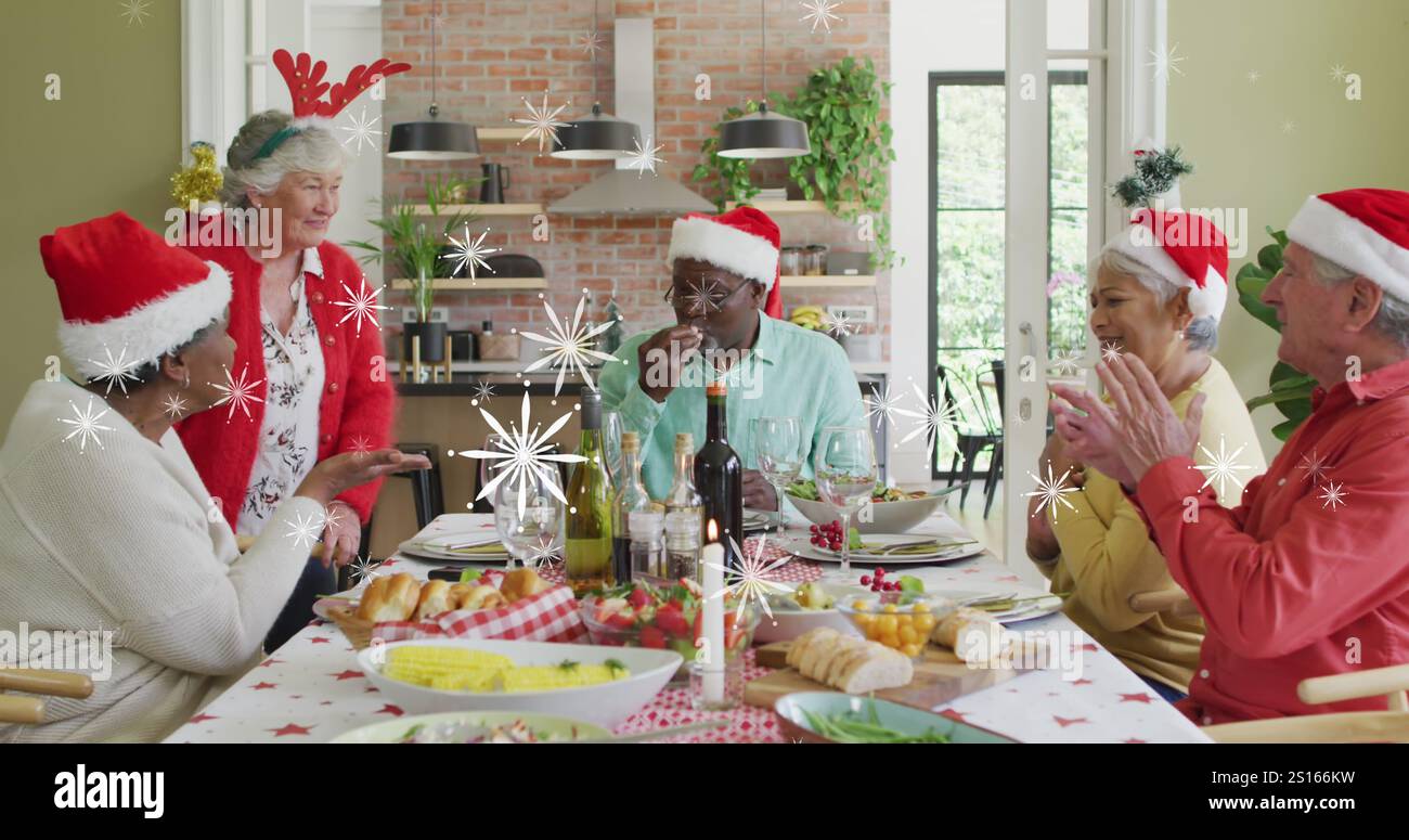 Image de neige tombant sur divers amis seniors dans des chapeaux de père noël à la table de repas de noël Banque D'Images