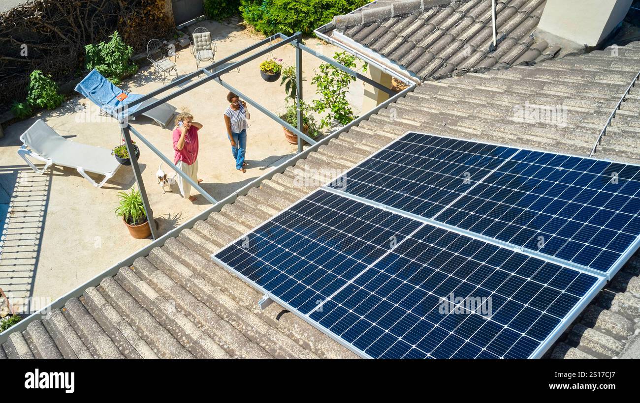 Une vue aérienne de deux femmes debout dans une cour arrière, célébrant l'installation de panneaux solaires sur le toit. La scène capture un moment de fierté Banque D'Images