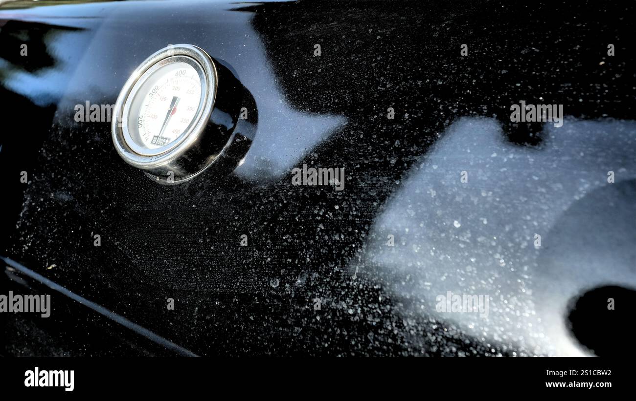 Indicateur de température circulaire sur un couvercle de gril extérieur de marque Weber avec lecture en degrés Celsius et Fahrenheit ; émail noir avec taches de saleté après la pluie Banque D'Images