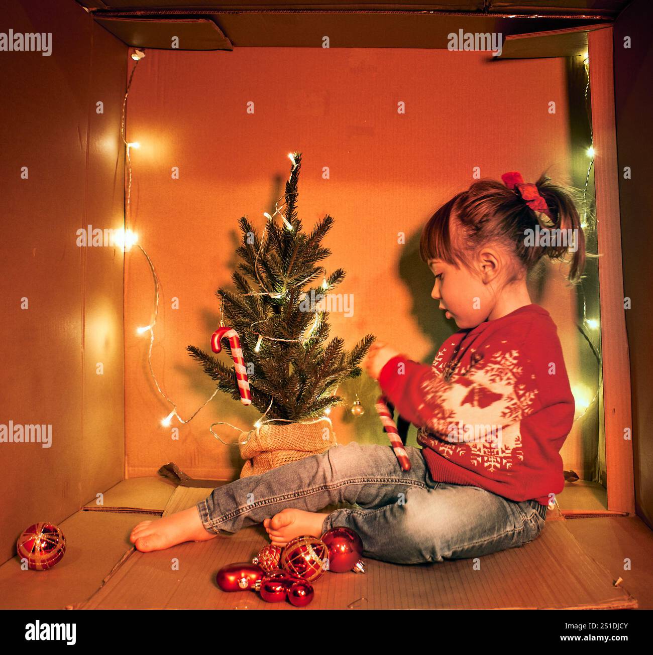 Un enfant dans un pull rouge avec des motifs blancs est assis dans une boîte en carton décorée de guirlandes lumineuses, tenant une canne à bonbons. Un petit sapin de Noël Banque D'Images