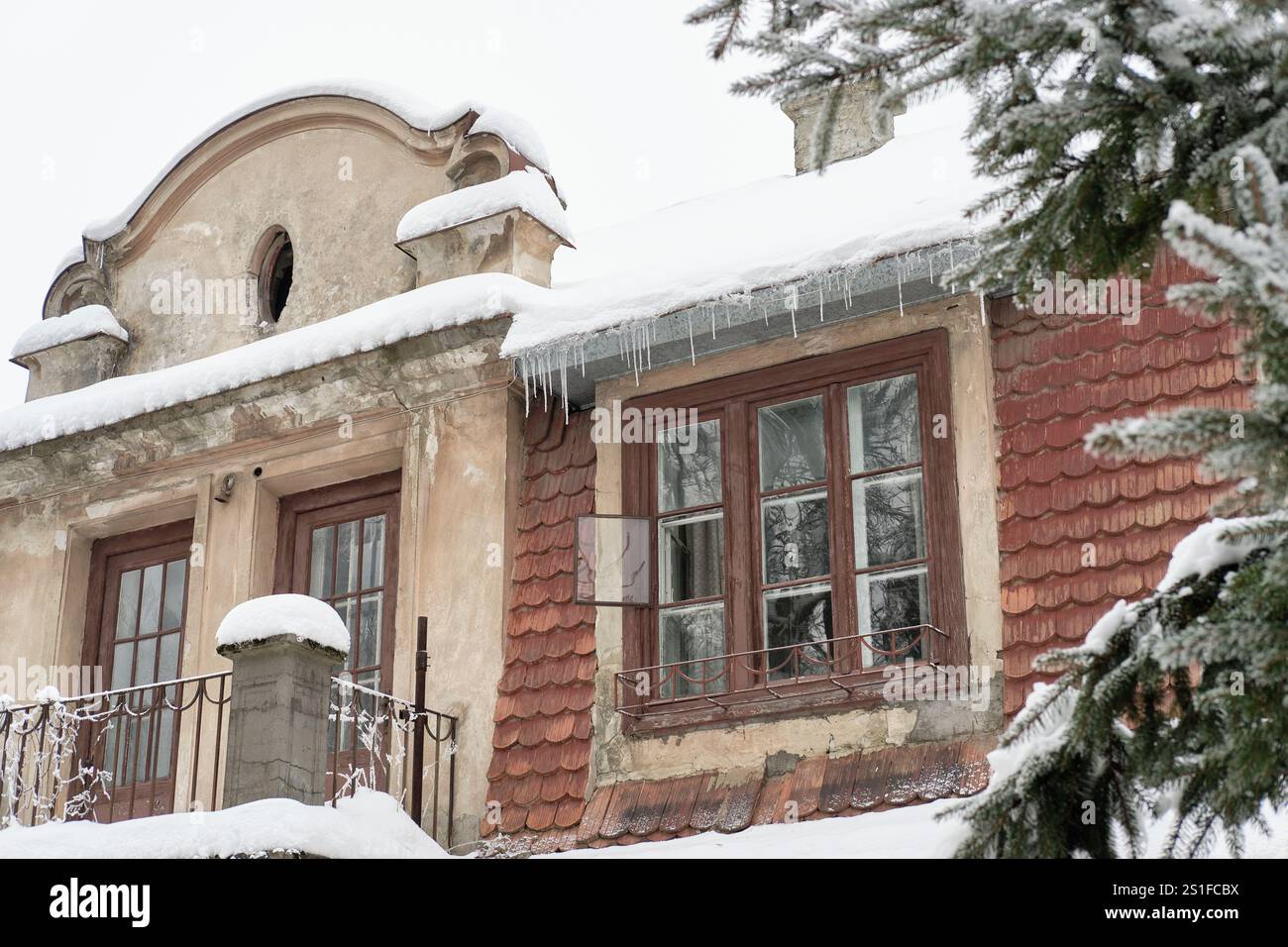 Ancien bâtiment rustique avec toit enneigé, glaçons suspendus à l'avant-toit et façade carrelée rouge. Une scène hivernale sereine mettant en valeur l'architecture historique A. Banque D'Images