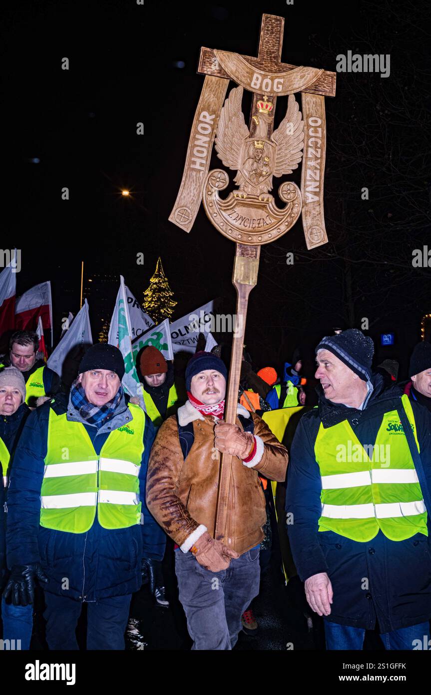 Un manifestant porte un bouclier attaché à une croix en bois portant la devise militaire polonaise « Dieu, honneur, Patrie » pendant la manifestation. Dans l'après-midi du 3 janvier, lors de la cérémonie de début de la présidence polonaise de l'UE, des agriculteurs polonais et d'autres travailleurs agricoles se rassemblent à Varsovie pour protester. Alors qu’Ursula von der Leyen se rend en Pologne pour la célébration du gala d’ouverture au Théâtre National, les manifestants se réunissent au bâtiment de la Commission européenne et défilent dans les rues jusqu’au théâtre. Les manifestants ont 5 revendications : arrêter les importations alimentaires en provenance des pays du Mercosur, t Banque D'Images