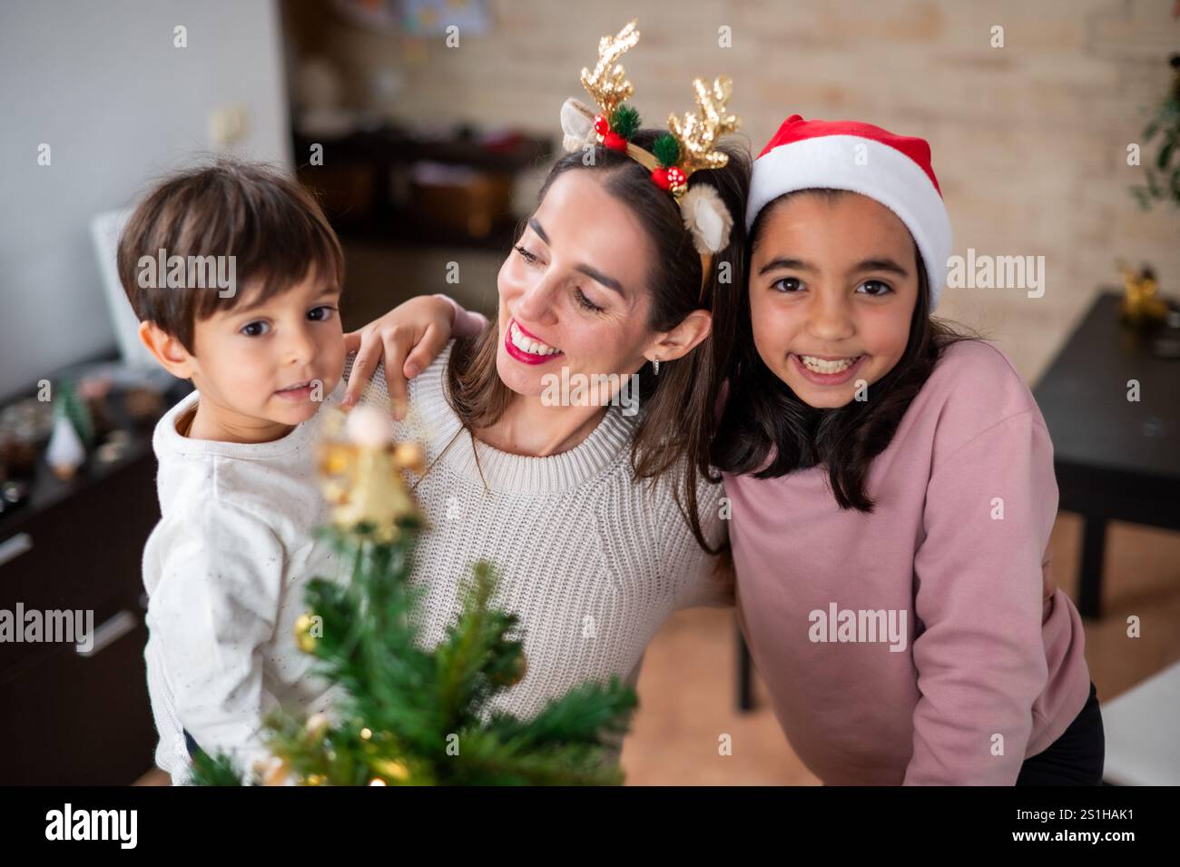 Une femme tient un petit sapin de Noël avec ses deux enfants. Les enfants portent des chapeaux de Père Noël et la femme porte un bandeau de cerf. Scène i. Banque D'Images