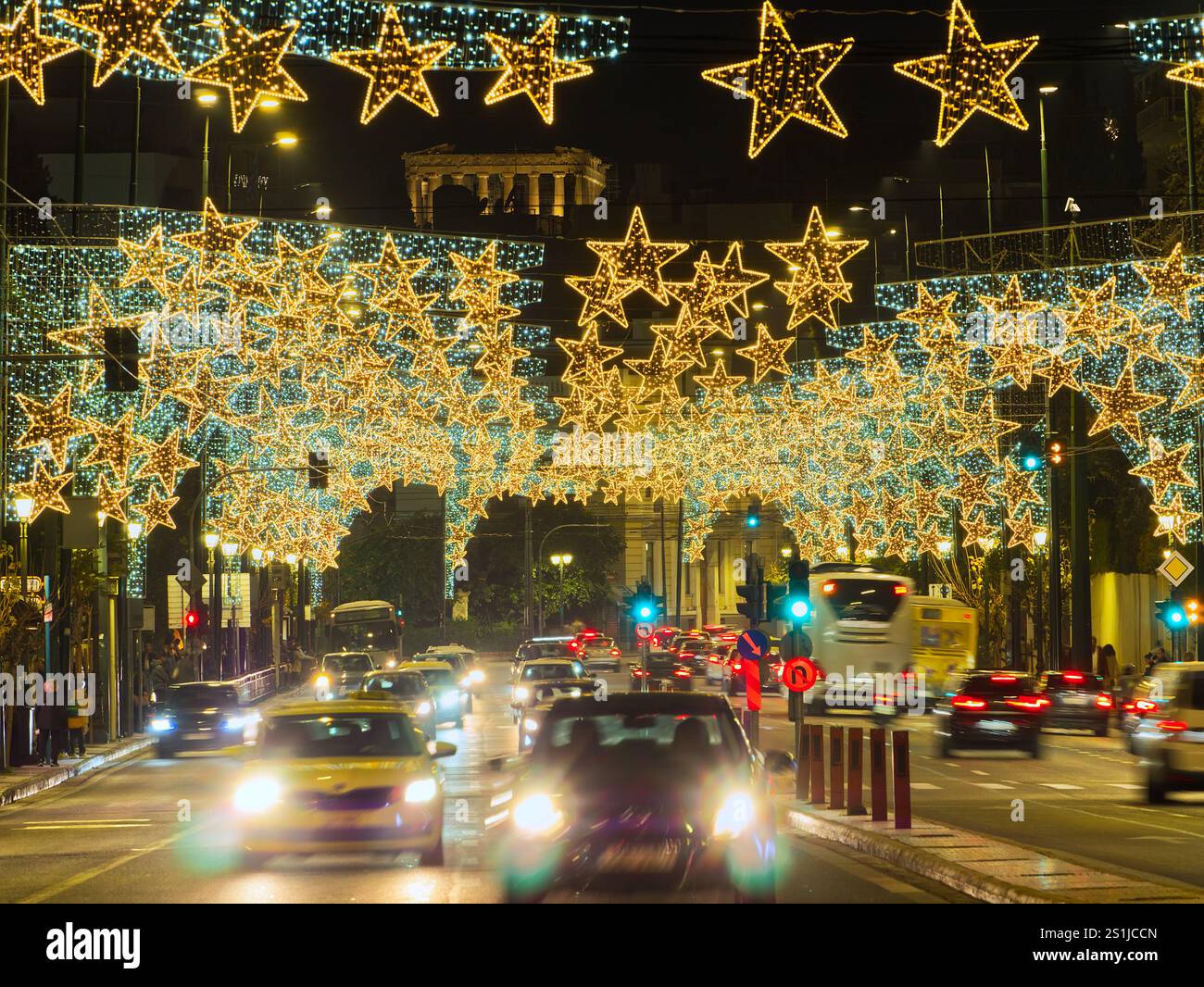 Athènes, Grèce - 3 janvier 2025 : rue urbaine illuminée par des lumières en forme d'étoile dorée et la circulation pendant la saison des fêtes. Acropole en vue. Banque D'Images