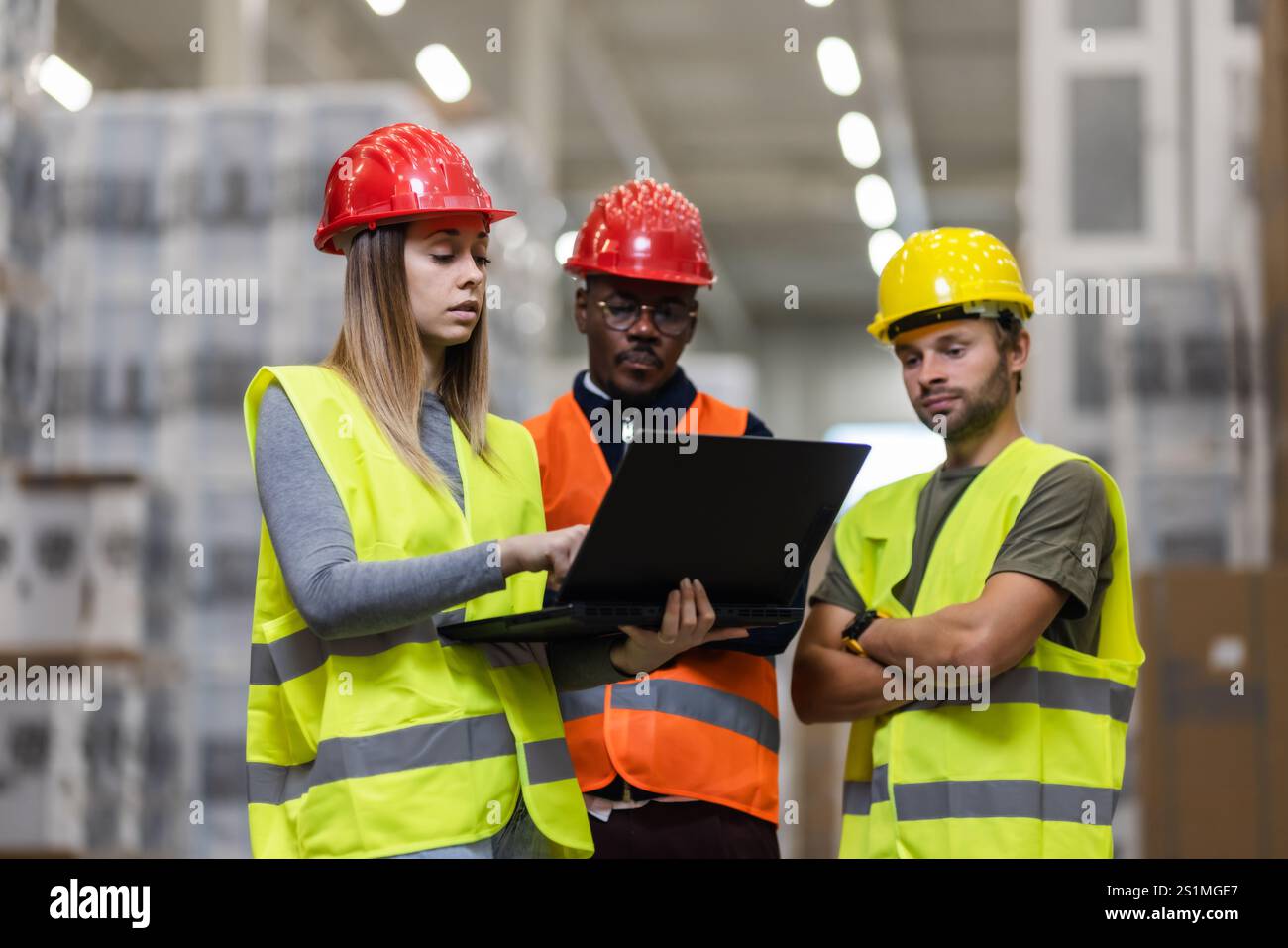 Les employés de l'entrepôt en équipement de sécurité collaborent autour d'un ordinateur portable, garantissant l'exactitude des stocks. Ils se concentrent sur l'efficacité, portant des casques de sécurité et des gilets, et Banque D'Images