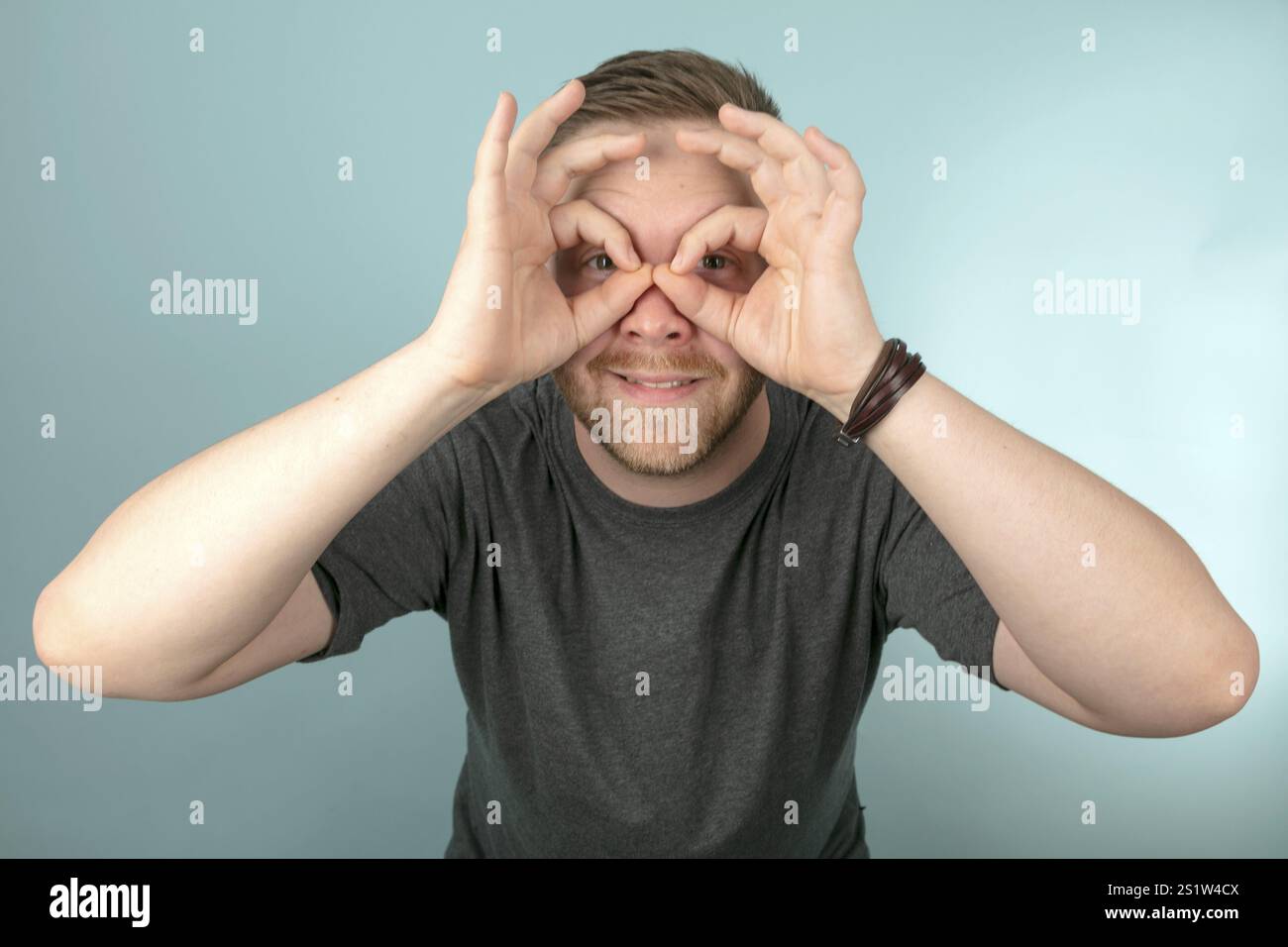 Junger Mann, der mit seinen Fingern eine Brillengeste um Seine Augen macht, waehrend er in einem Studioportraet in die Kamera blickt Banque D'Images