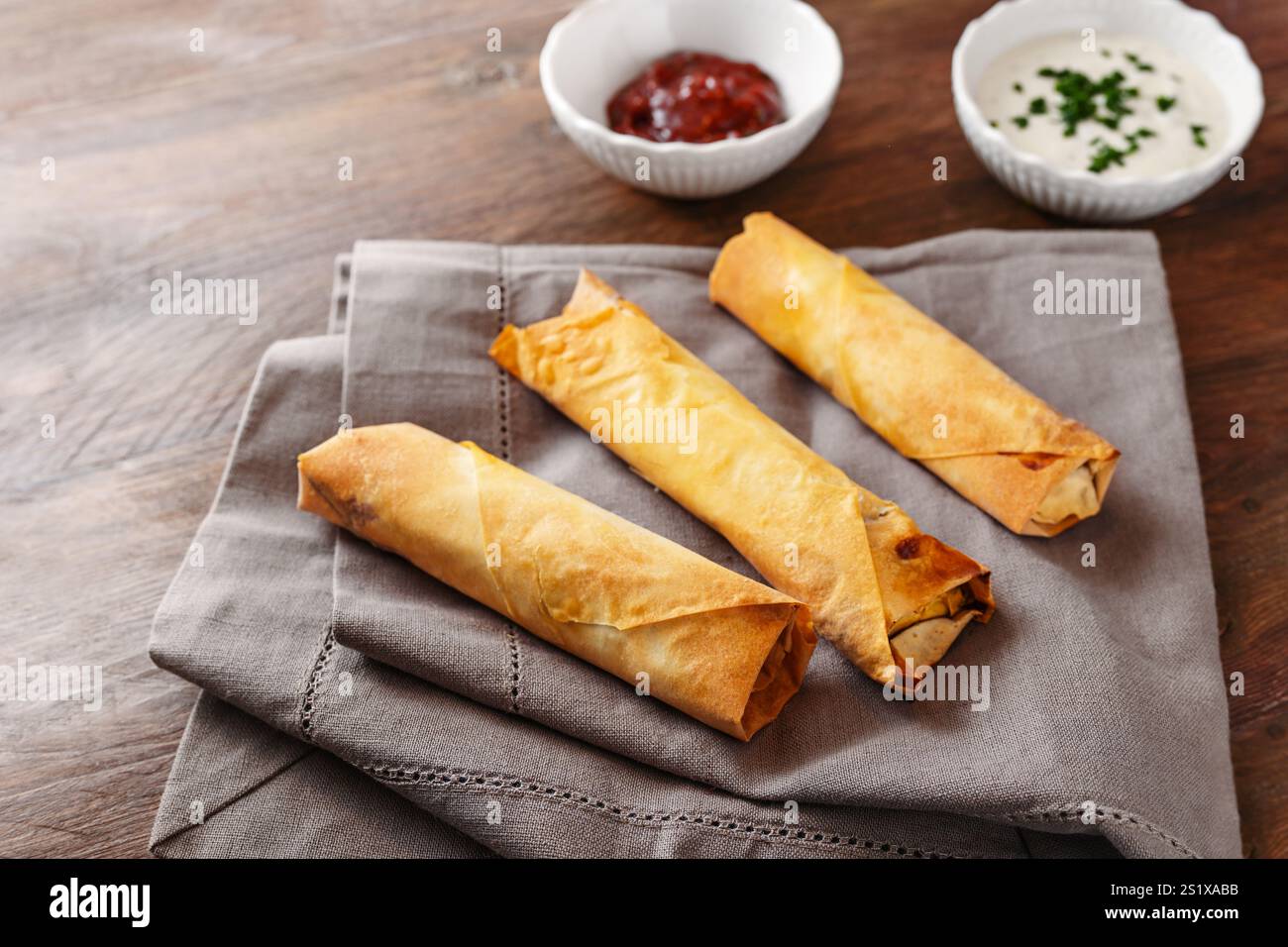 Trois rouleaux de pâtisserie filo croustillants cuits au four ou rouleaux de printemps avec deux trempettes sur une serviette grise et une table en bois rustique, nourriture maison, espace copie, selec Banque D'Images