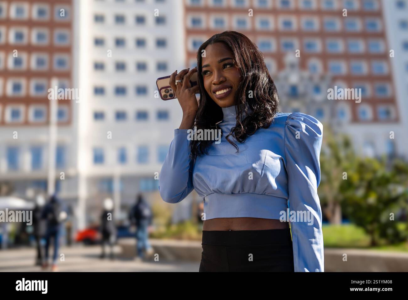 Femme d'affaires souriante écoutant un message vocal sur son smartphone tout en marchant dans une ville moderne Banque D'Images