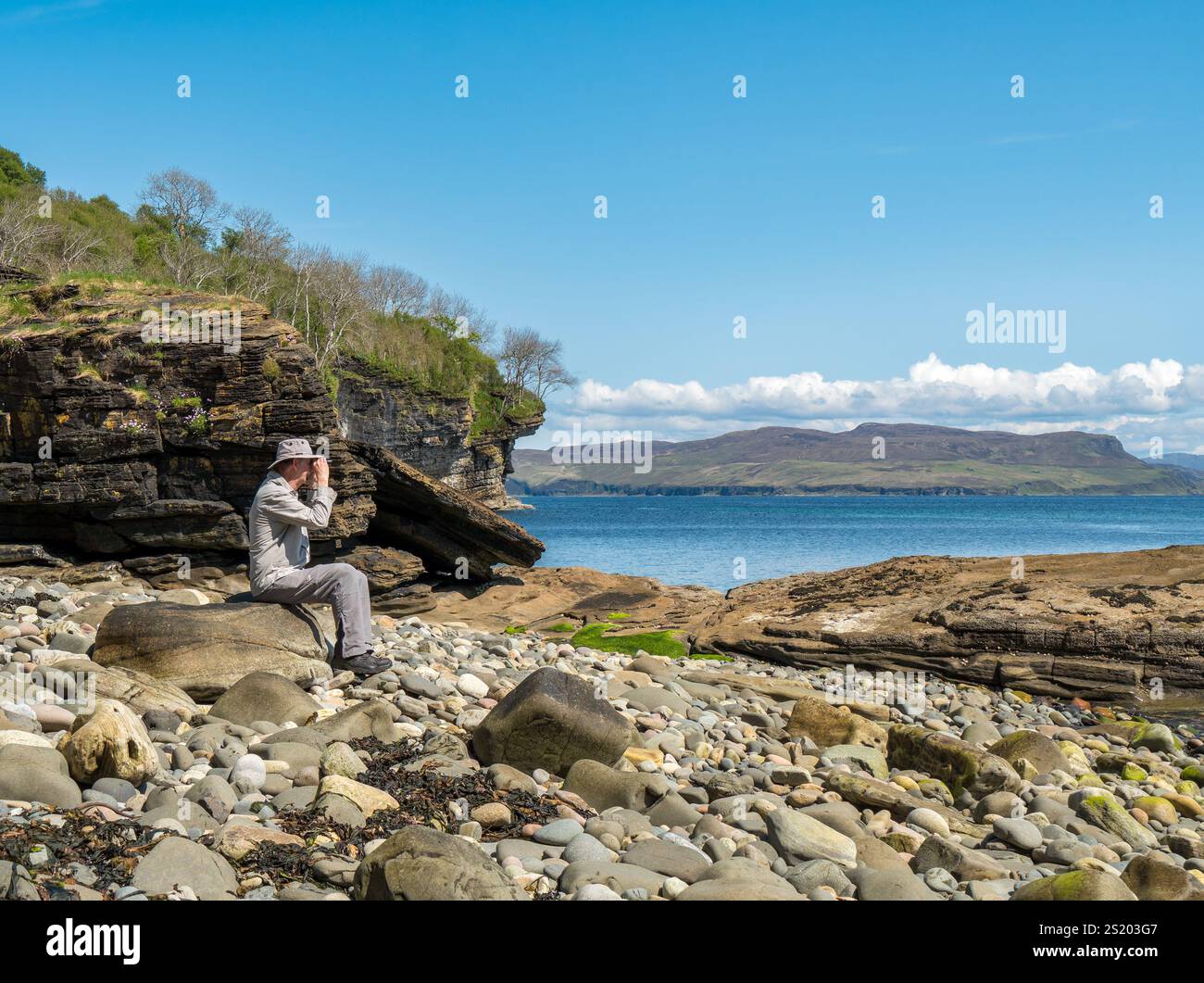 Homme adulte observant la faune à travers des jumelles sur une plage rocheuse par une journée ensoleillée en mai, Glasnakille près d'Elgol, île de Skye, Écosse, Royaume-Uni Banque D'Images