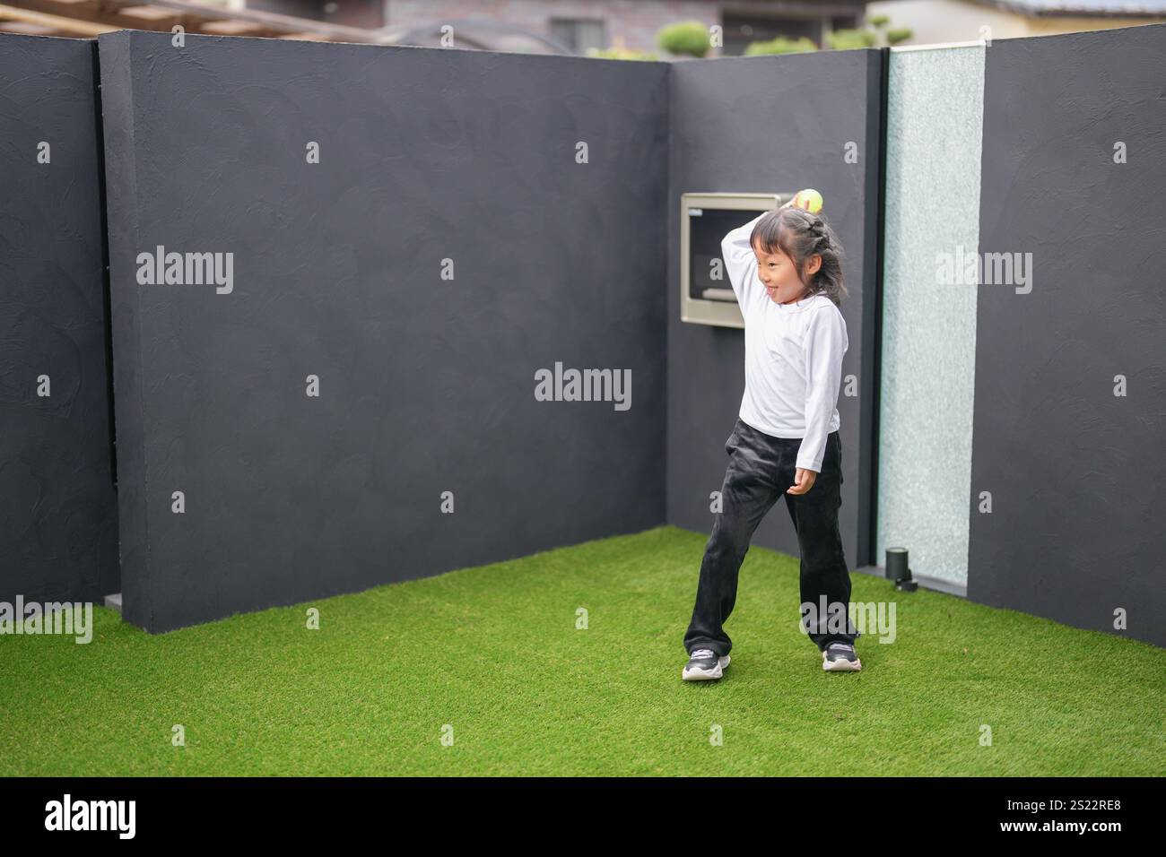 Un parent japonais et un enfant, un homme dans la vingtaine et une fille de 6 ans, jouent en se jetant des balles en plastique vert l'un sur l'autre dans le jardin clôturé de Banque D'Images