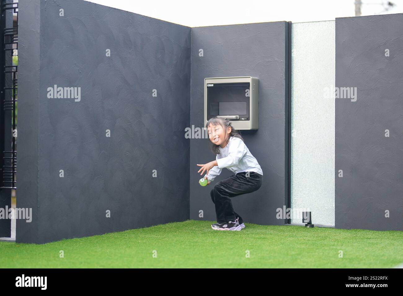 Un parent japonais et un enfant, un homme dans la vingtaine et une fille de 6 ans, jouent en se jetant des balles en plastique vert l'un sur l'autre dans le jardin clôturé de Banque D'Images
