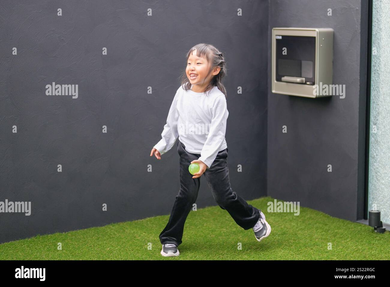 Un parent japonais et un enfant, un homme dans la vingtaine et une fille de 6 ans, jouent en se jetant des balles en plastique vert l'un sur l'autre dans le jardin clôturé de Banque D'Images