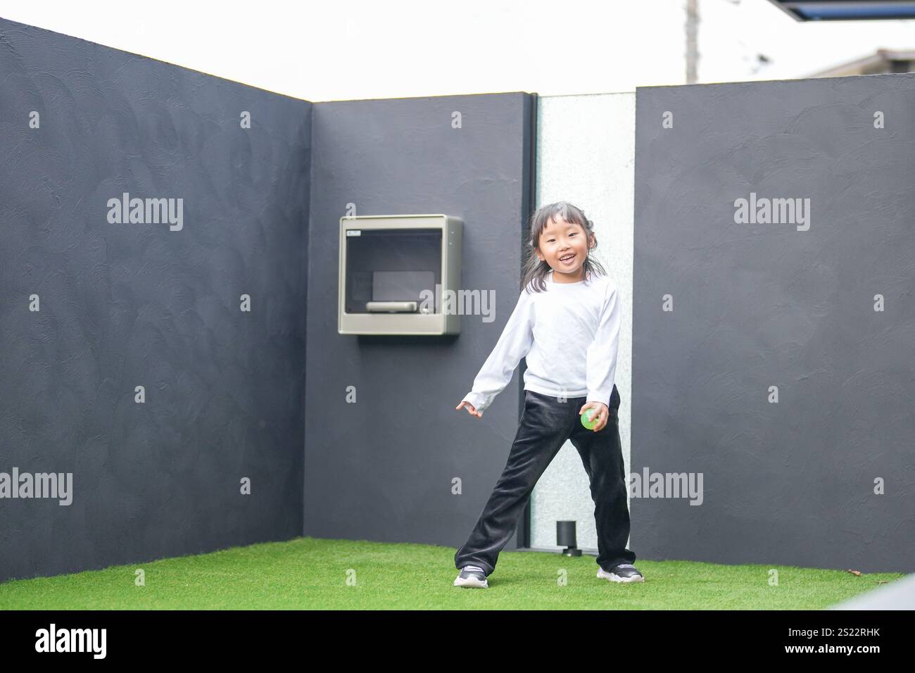 Un parent japonais et un enfant, un homme dans la vingtaine et une fille de 6 ans, jouent en se jetant des balles en plastique vert l'un sur l'autre dans le jardin clôturé de Banque D'Images