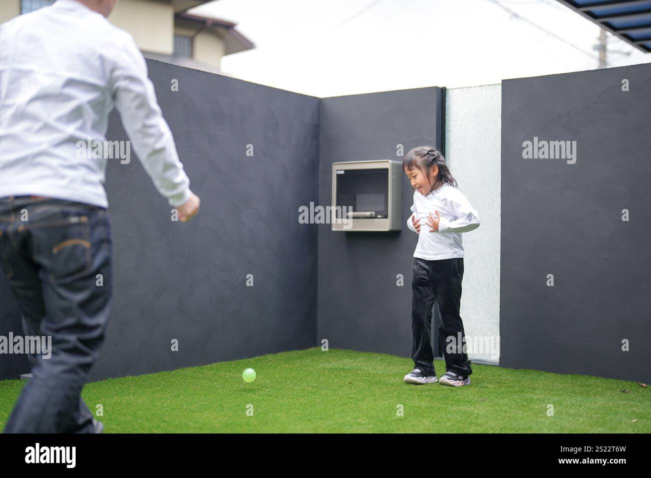 Un parent japonais et un enfant, un homme dans la vingtaine et une fille de 6 ans, jouent en se jetant des balles en plastique vert l'un sur l'autre dans le jardin clôturé de Banque D'Images