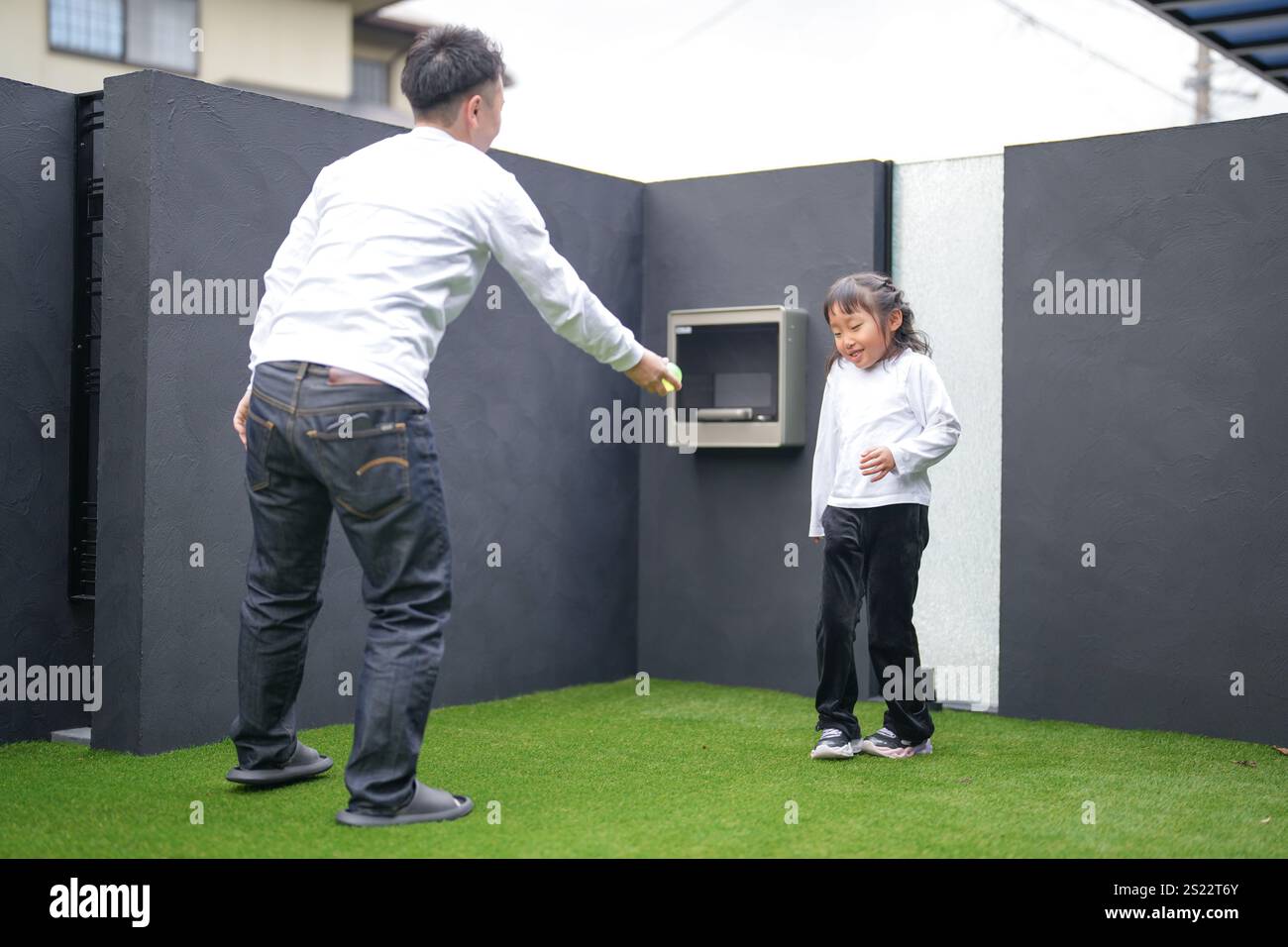 Un parent japonais et un enfant, un homme dans la vingtaine et une fille de 6 ans, jouent en se jetant des balles en plastique vert l'un sur l'autre dans le jardin clôturé de Banque D'Images