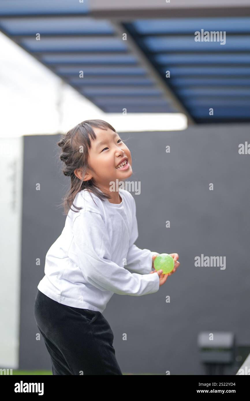 Un parent japonais et un enfant, un homme dans la vingtaine et une fille de 6 ans, jouent en se jetant des balles en plastique vert l'un sur l'autre dans le jardin clôturé de Banque D'Images