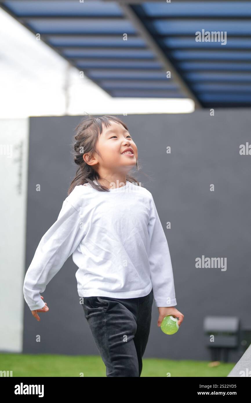 Un parent japonais et un enfant, un homme dans la vingtaine et une fille de 6 ans, jouent en se jetant des balles en plastique vert l'un sur l'autre dans le jardin clôturé de Banque D'Images