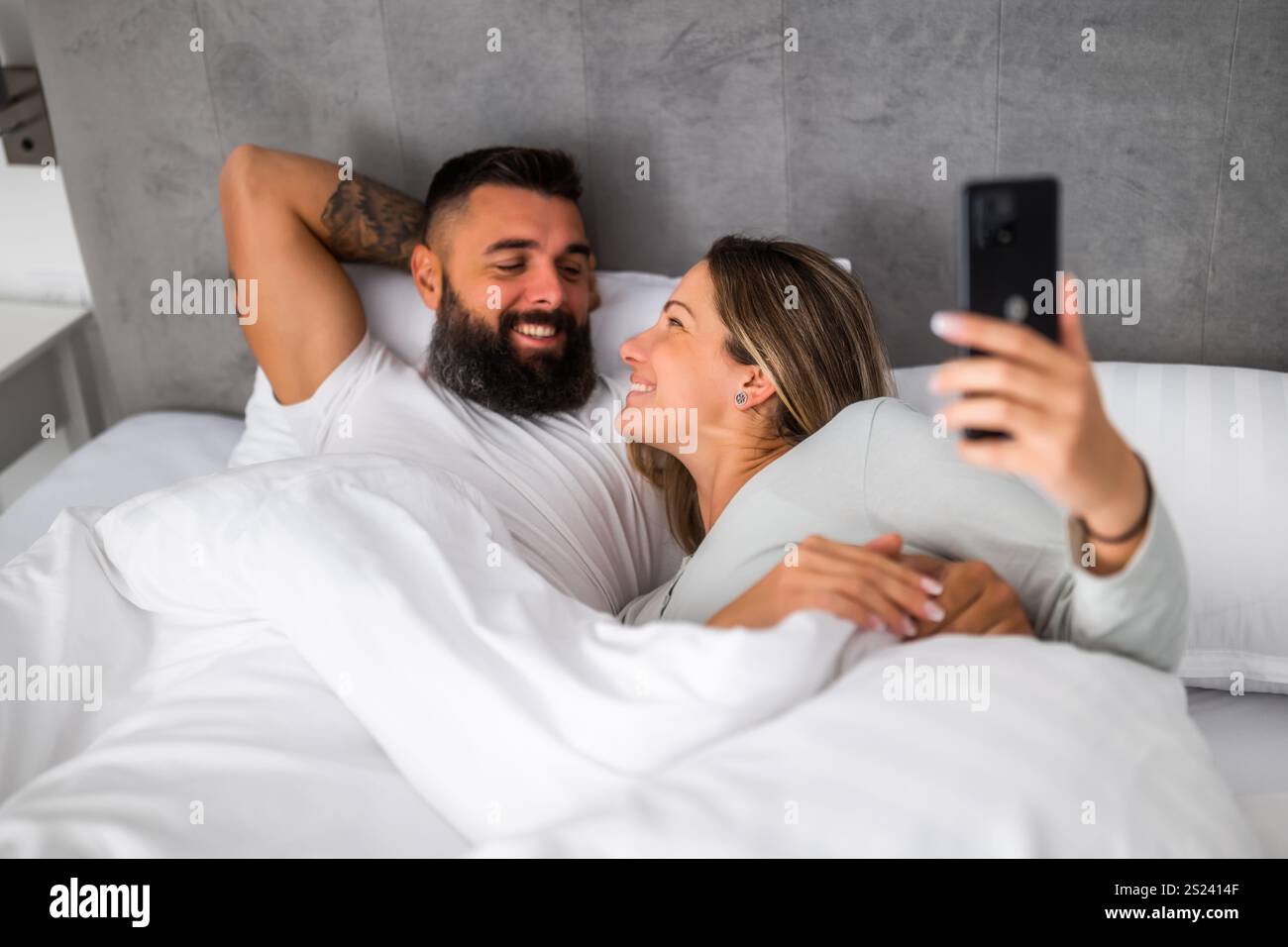 Jeune couple aimant heureux est couché dans le lit le matin. Ils sont relaxants et utilisent un smartphone. Banque D'Images