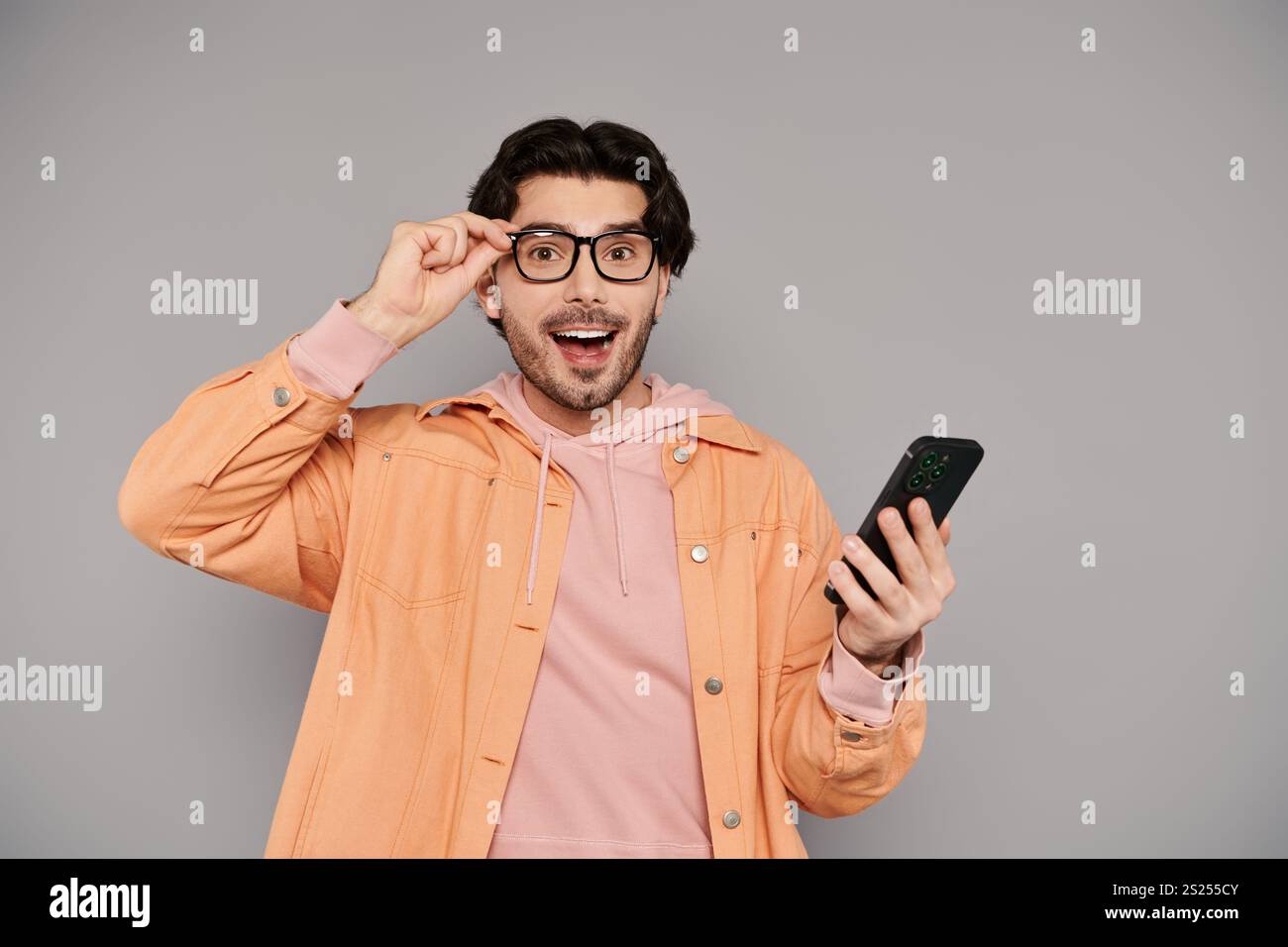 Un jeune homme beau aux cheveux bruns montre de la joie et de la surprise en utilisant son smartphone. Banque D'Images