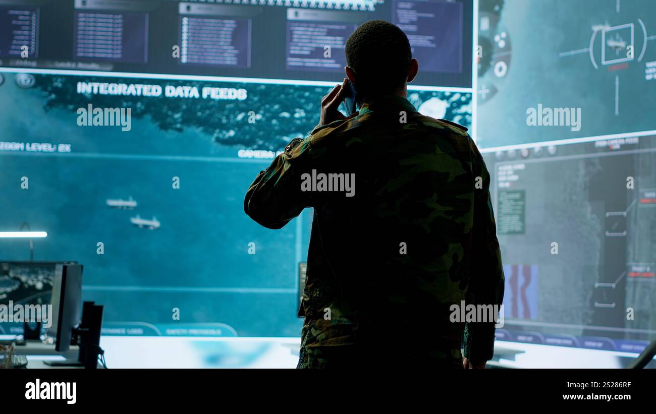 Un militaire répond à un appel téléphonique dans un centre de contrôle de mission, travaillant avec une technologie de pointe et des images satellites pour suivre les menaces potentielles et les données en temps réel. Activités de renseignement. Caméra A. Banque D'Images