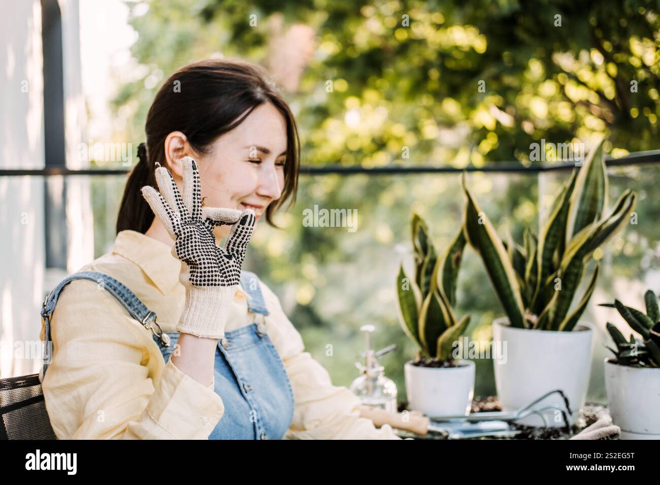 Une femme en gants fait des gestes avec un signe OK entourée de plantes en pot sur un balcon ensoleillé. Représente le bonheur et l'accomplissement du jardinage. Banque D'Images