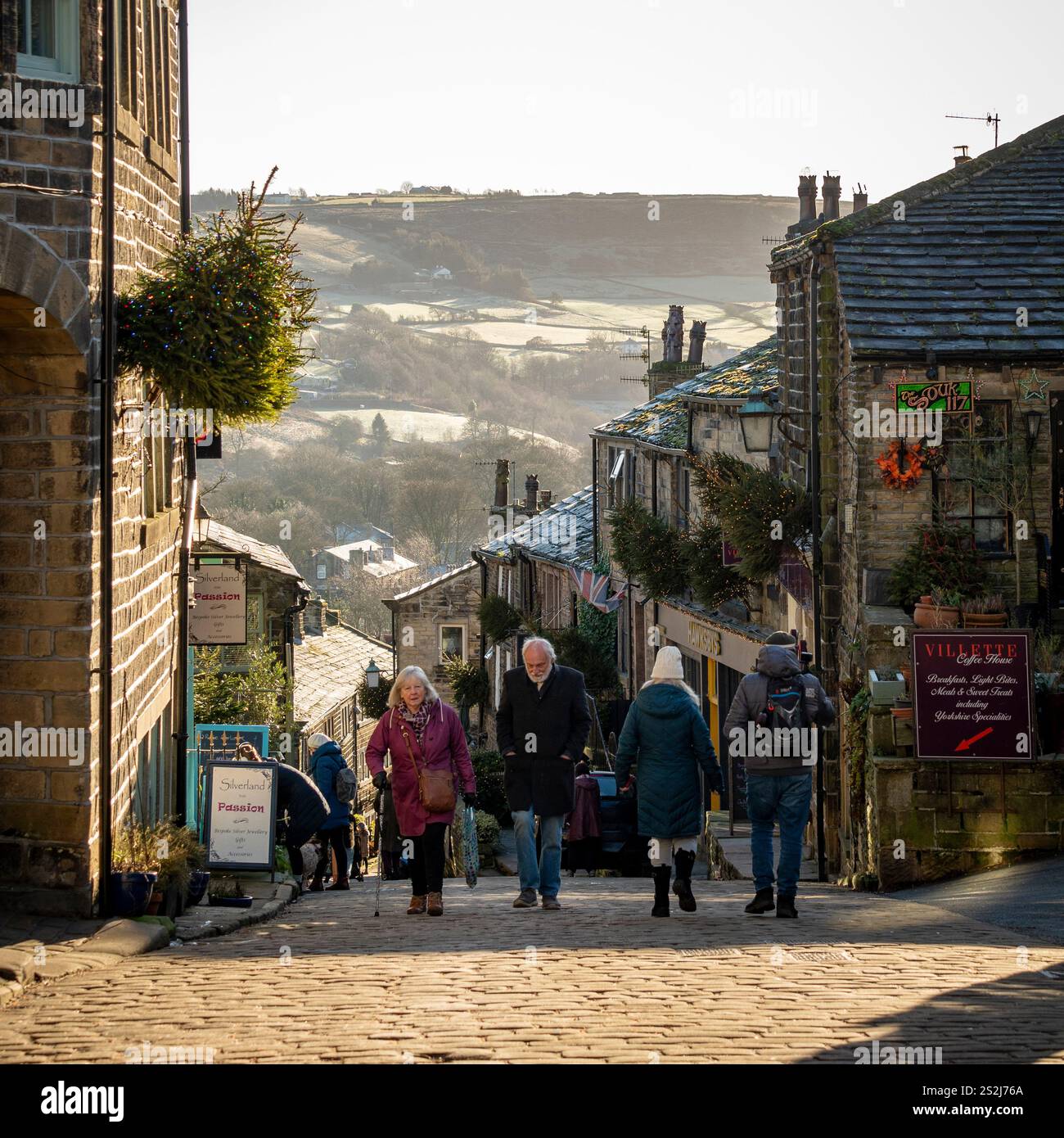 Les touristes se promènent dans la rue principale escarpée de Haworth, avec les landes pittoresques qui s'étendent au loin par une journée d'hiver ensoleillée. Banque D'Images