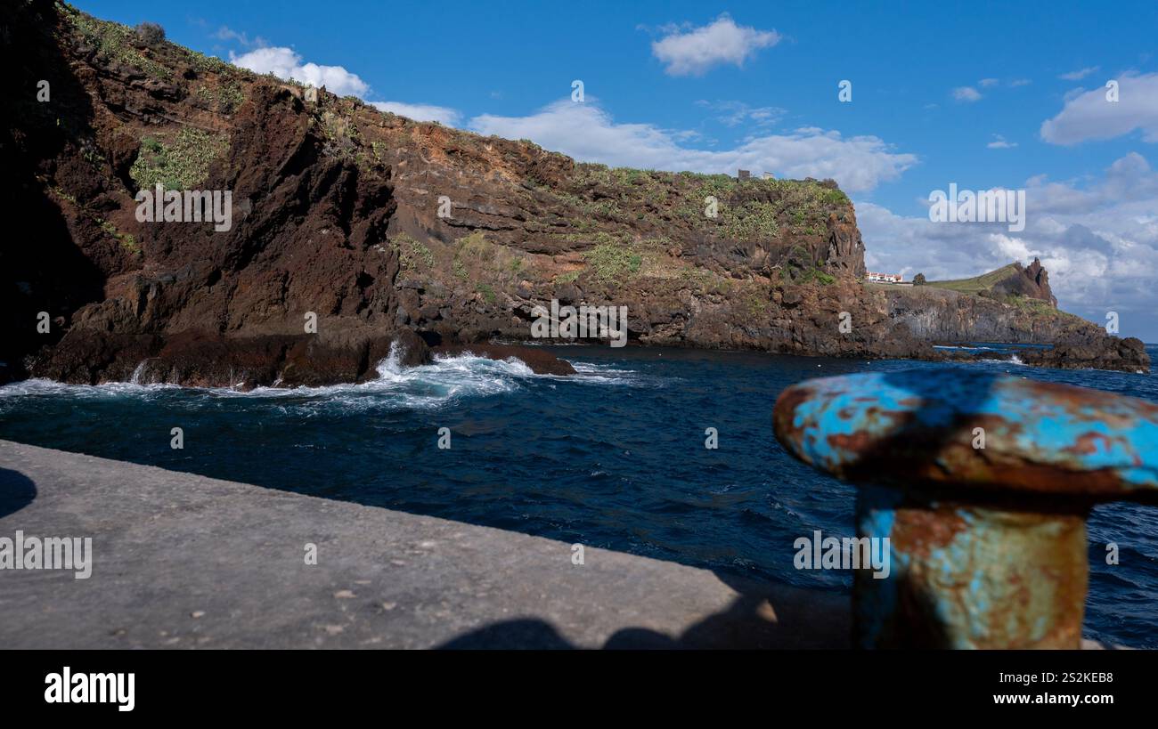 Une scène côtière avec des falaises escarpées couvertes de verdure, des vagues s'écrasant sur les rochers, et un poste d'amarrage rouillé au premier plan sous un ciel ensoleillé. Banque D'Images