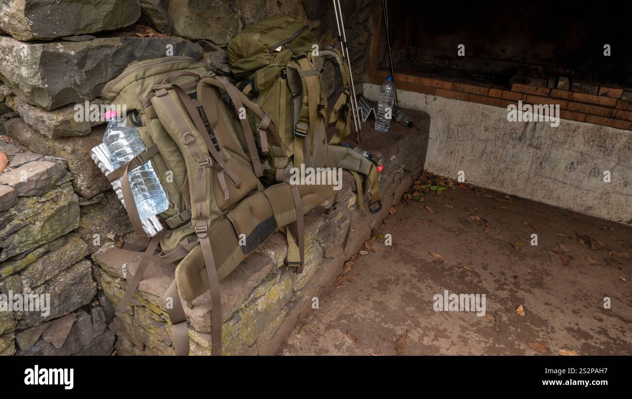 Sacs à dos de randonnée avec des bouteilles d'eau reposant sur un banc de pierre dans un abri rustique, entouré d'équipements de plein air et d'un environnement altéré. Banque D'Images Sacs à dos de randonnée avec des bouteilles d'eau reposant sur un banc de pierre dans un abri rustique, entouré d'équipements de plein air et d'un environnement altéré. Banque D'Images