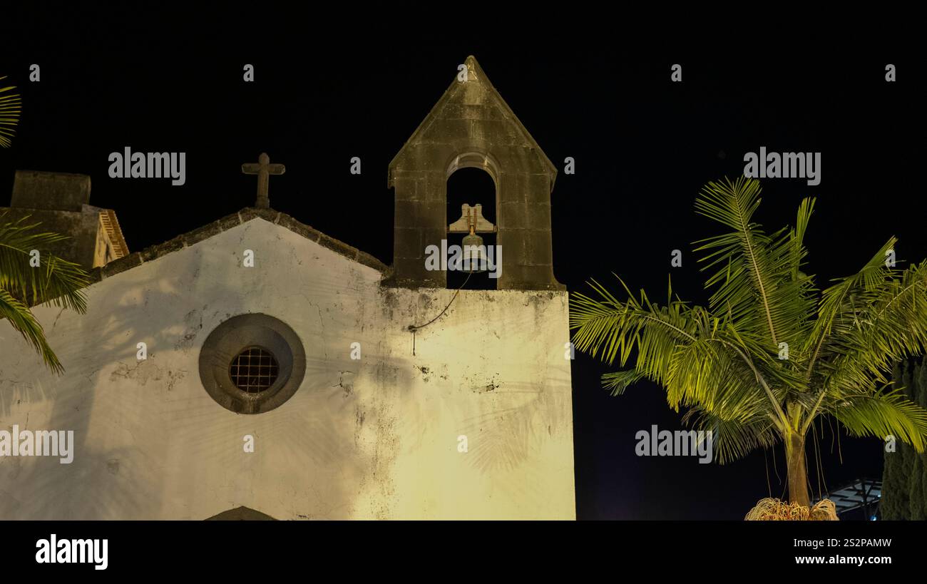 Une petite église historique avec un clocher en pierre illuminé la nuit, encadré par des palmiers, sous un ciel sombre, créant une atmosphère sereine. Banque D'Images