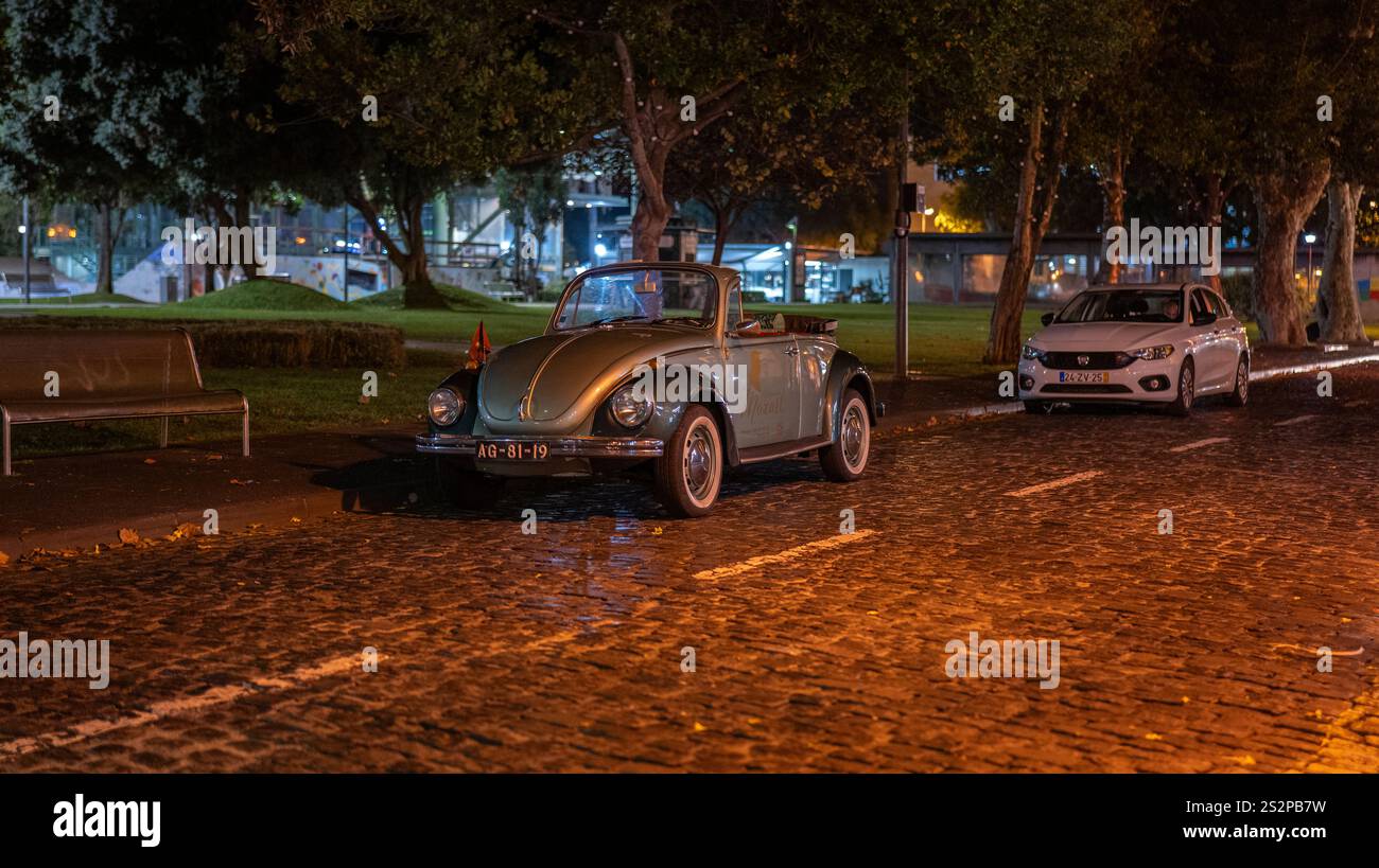 Une voiture cabriolet vintage garée sur une rue pavée humide la nuit, éclairée par des lampadaires chauds, avec des arbres et un cadre urbain calme dans le b. Banque D'Images