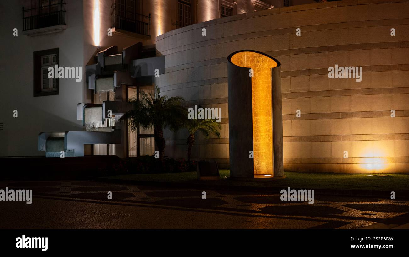 Une sculpture urbaine moderne illuminée par un éclairage chaleureux la nuit, posée contre une façade de bâtiment courbe et entourée de palmiers et architecturale Banque D'Images