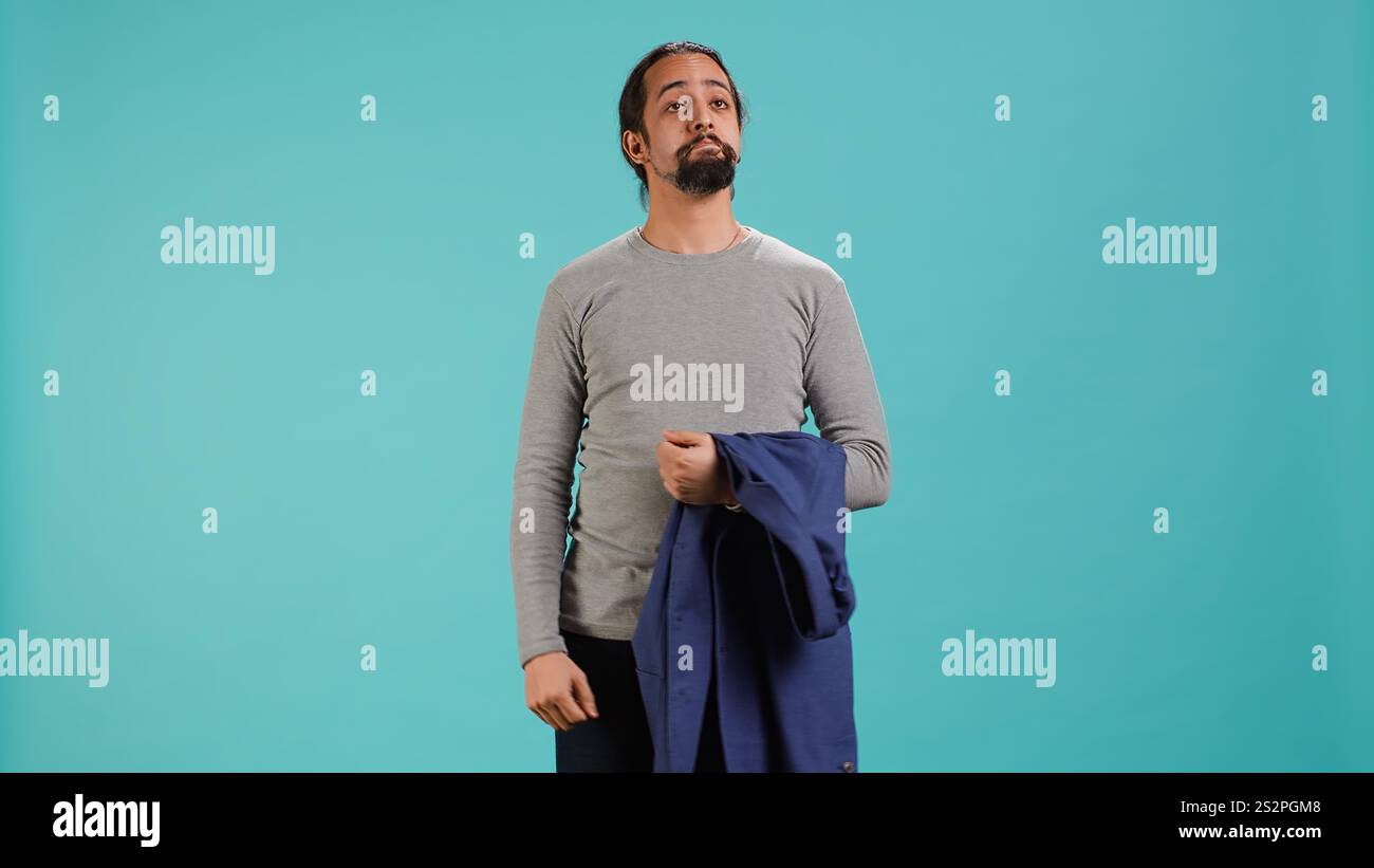 Homme tenant la veste dans une main tout en regardant autour, contemplant, créant un sentiment de curiosité. Personne tenant le manteau montrant la curiosité pour l'environnement, fond de studio, caméra A. Banque D'Images