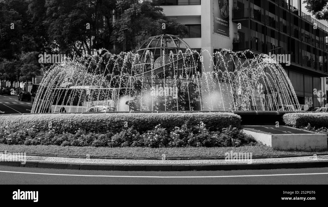 Photographie en noir et blanc d'une fontaine circulaire entourée de haies taillées et de fleurs dans un cadre urbain. La fontaine présente des ruisseaux voûtés Banque D'Images