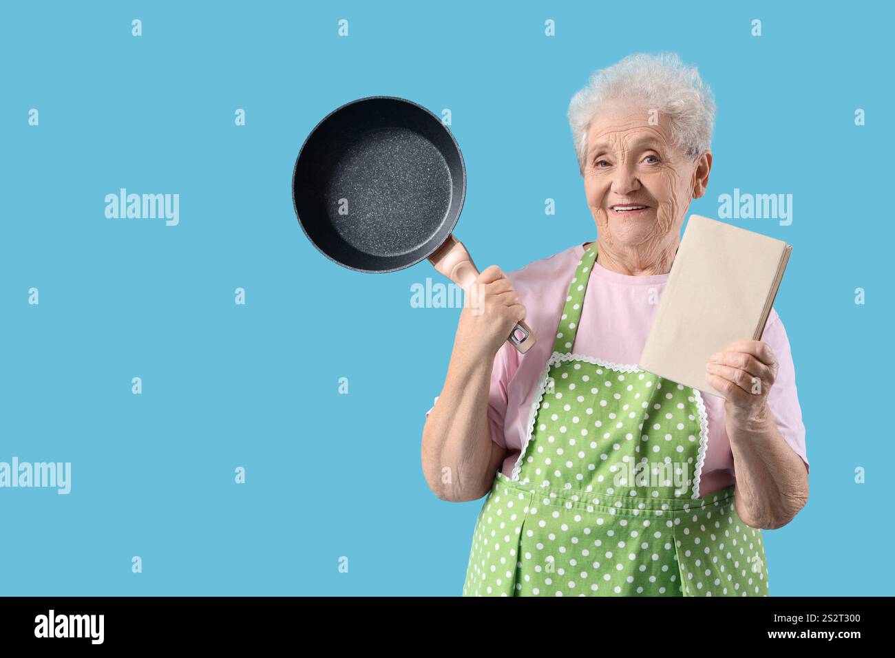 Femme senior dans le tablier avec poêle à frire et livre de recettes sur fond bleu Banque D'Images