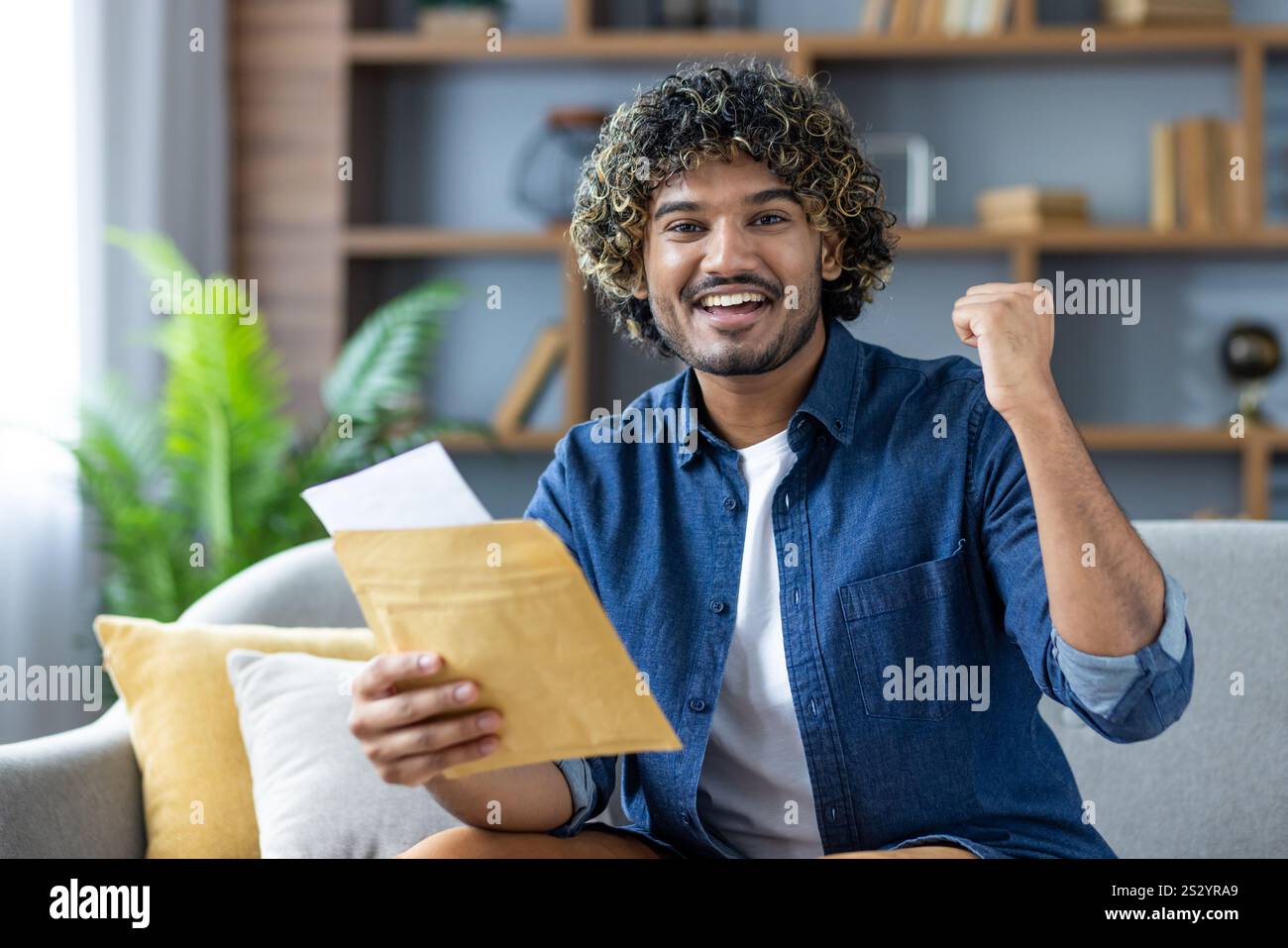 Gagnant de bonnes nouvelles, homme avec l'enveloppe de courrier regardant joyeusement la caméra, assis dans le salon à la maison, a reçu un message de notification par courrier sur de bons résultats. Banque D'Images