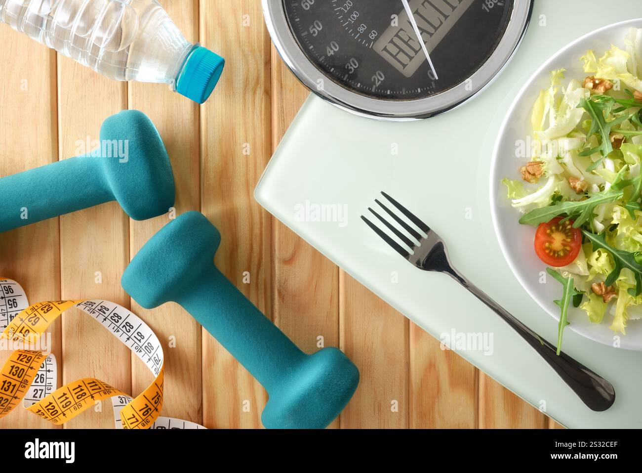 Détail du concept de régime de perte de poids avec assiette avec salade sur balance avec couverts sur un sommier à lattes en bois avec haltères, ruban à mesurer et bouteille de Banque D'Images