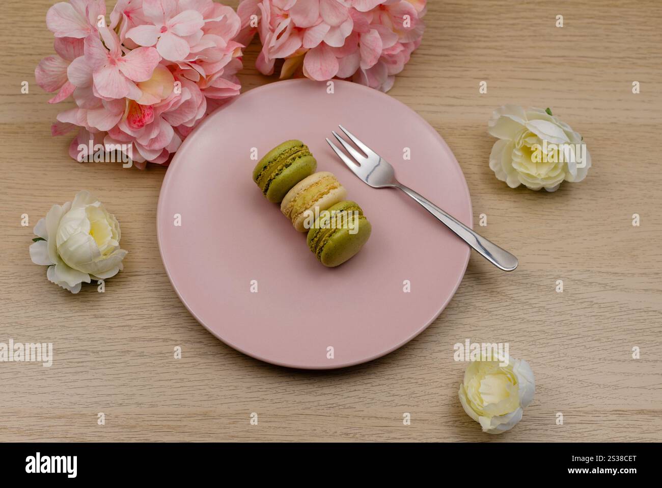 Macarons pistache et vanille sur une assiette rose sur une table en bois avec en arrière-plan des fleurs de couleur rose et crème Banque D'Images
