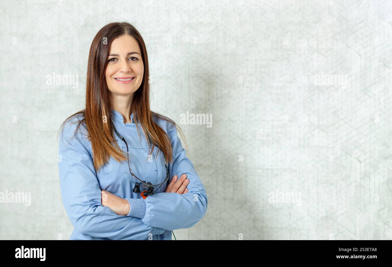 Dentiste confiante portant une loupe binoculaire souriant les bras croisés en clinique dentaire Banque D'Images