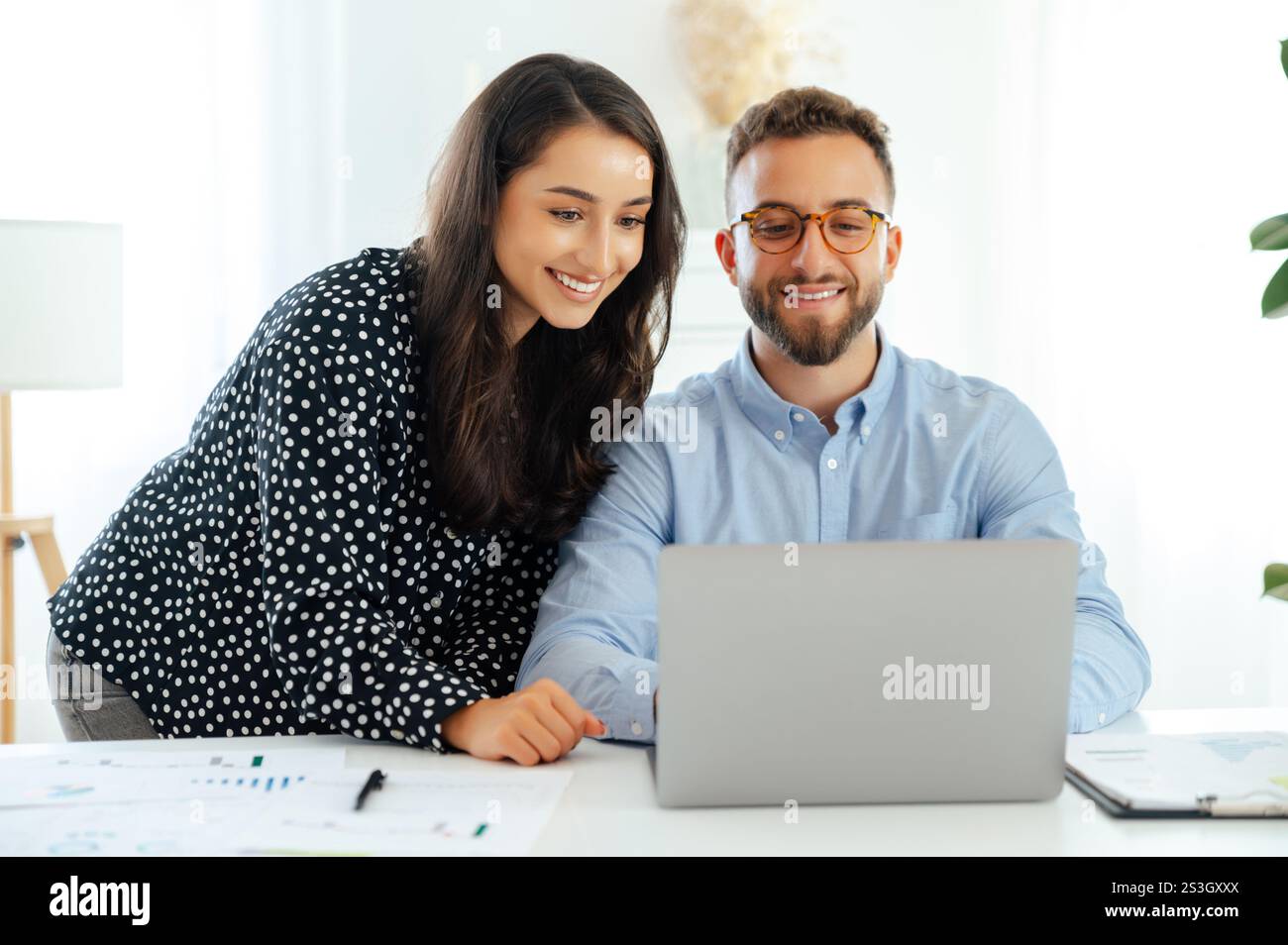 Concept de travail d'équipe. Femme souriante dans une chemise penchée sur un homme dans une chemise bleue, tous deux regardant un écran d'ordinateur portable, collaborant à un projet dans un cadre de bureau moderne lumineux avec des graphiques sur la table Banque D'Images