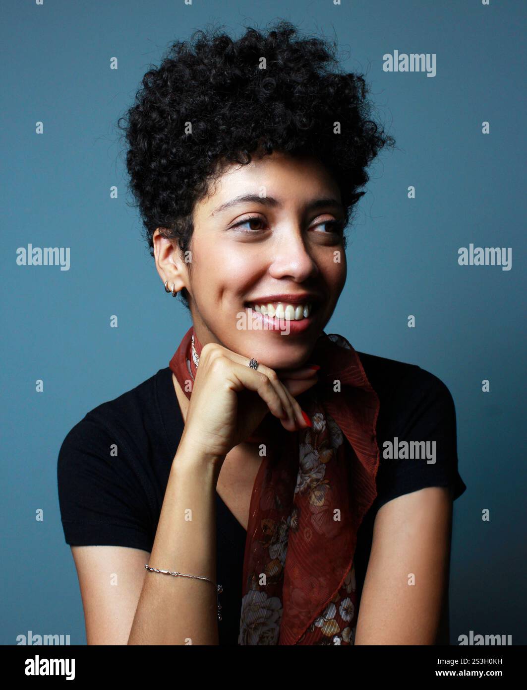 Portrait vertical de belle femme de peau brune souriante et regardant latéralement. Modèle féminin à cheveux bouclés courts. Femme afro-américaine avec le menton sur la main. Banque D'Images