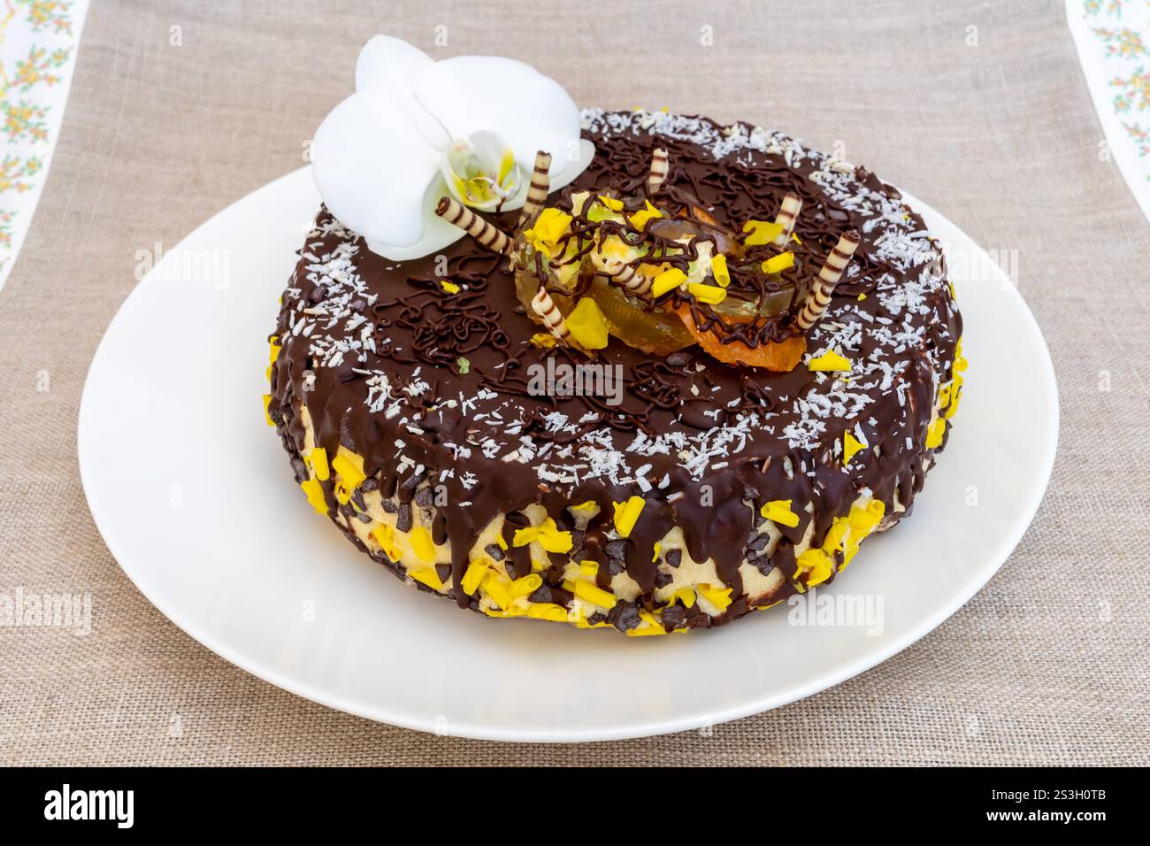 Un gâteau au chocolat joliment décoré repose sur une assiette blanche. Le gâteau présente des copeaux de chocolat, des accents de fruits tropicaux et un élégant orch blanc Banque D'Images
